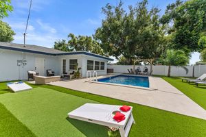 Backyard with pool, cornhole, and seating on green lawn. Blue sky, white house.