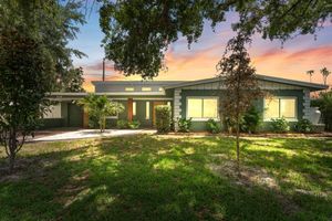 Green ranch-style house with manicured lawn, framed by trees under a colorful sunset sky.