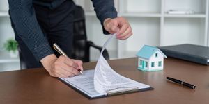 A person signing a document on a clipboard, next to a model house and pen on a desk.
