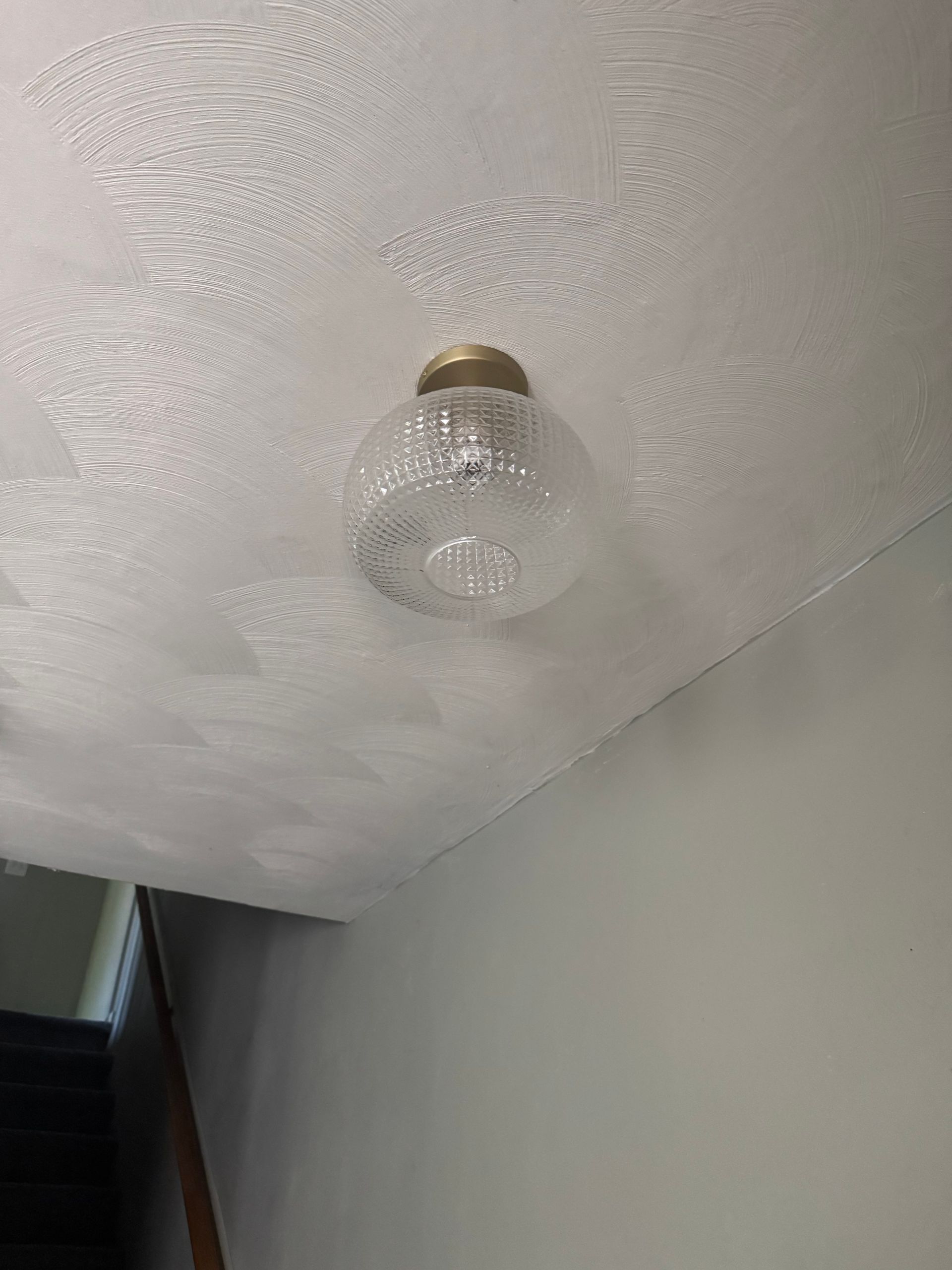 Clear glass ceiling light fixture on a white textured ceiling near a stairwell.