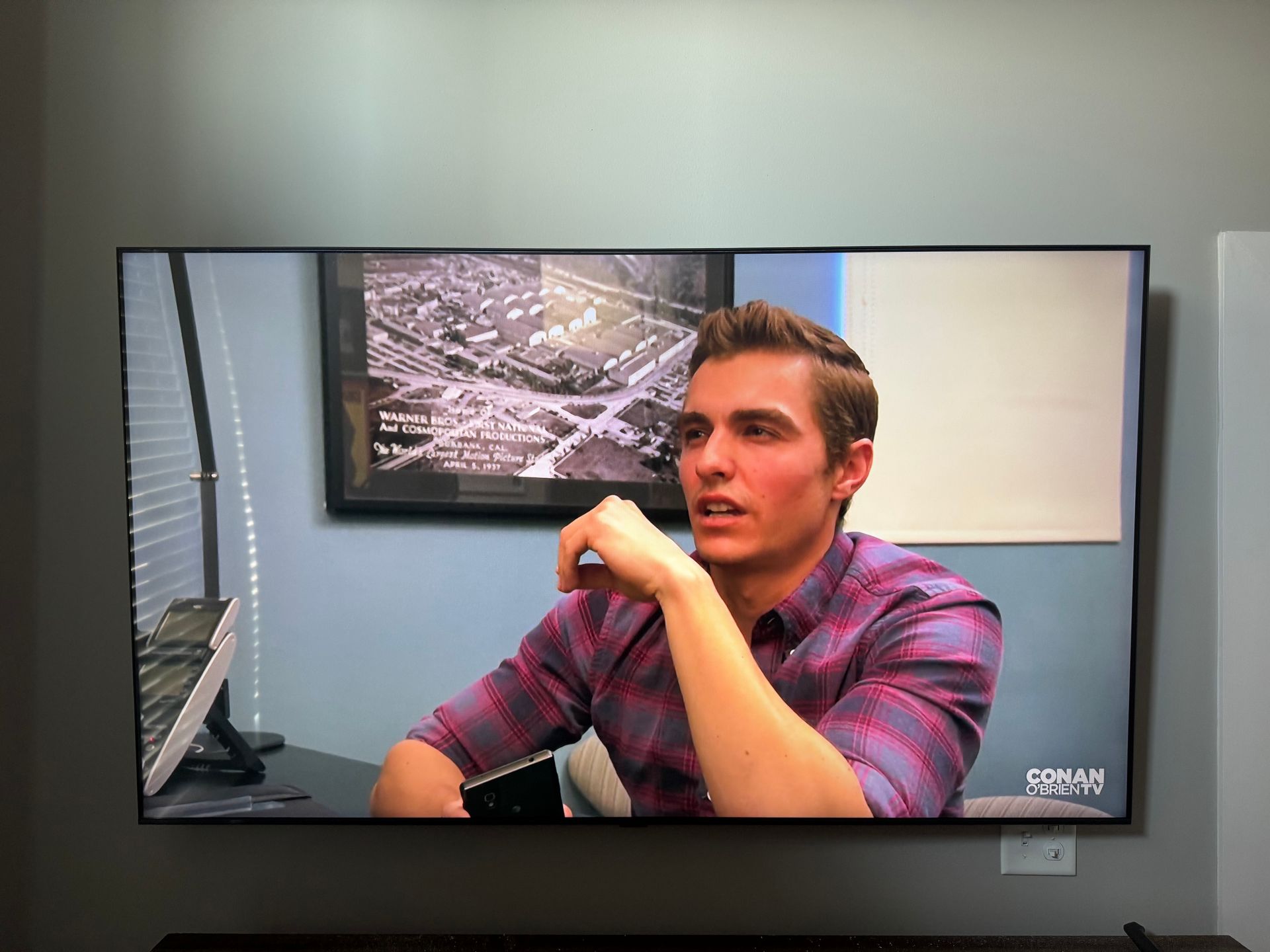 Man sitting at a desk in an office, leaning on his hand and looking at a computer monitor on the wall.