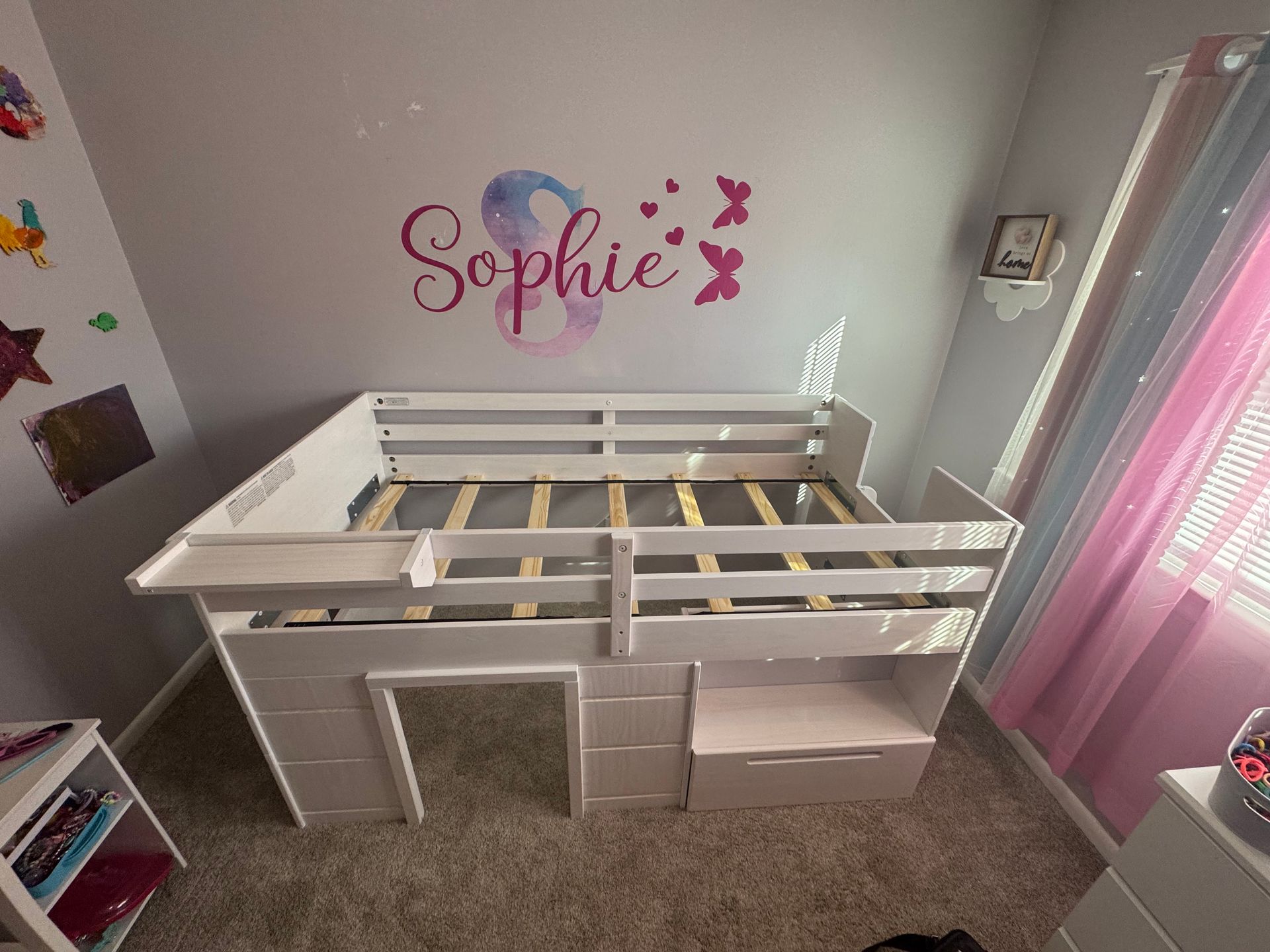 White loft bed frame in a child’s bedroom with pink curtains and a “Sophie” wall decal