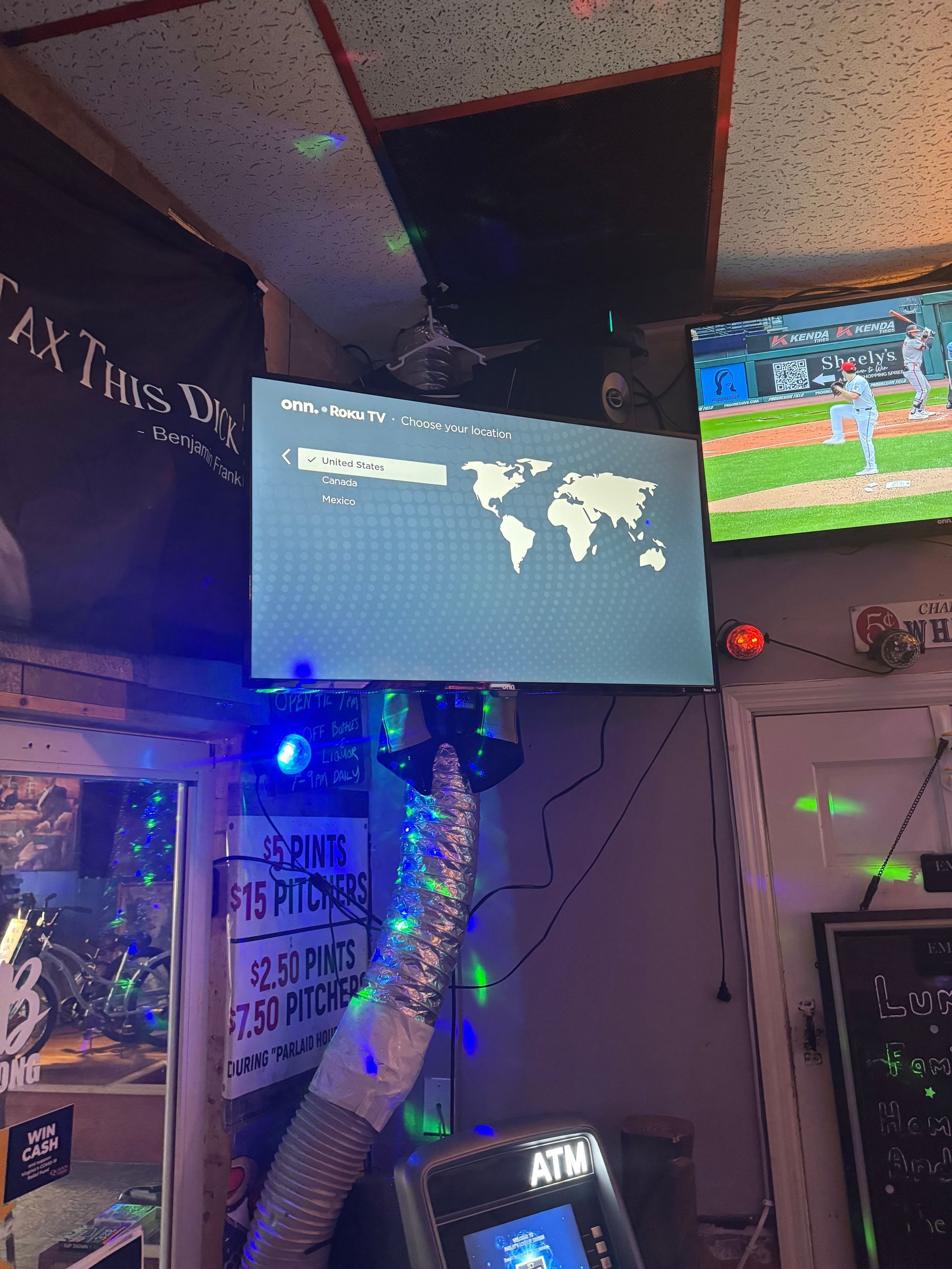 TV screen in a cluttered room shows a world map with text and graphics, beside a second TV displaying a football game