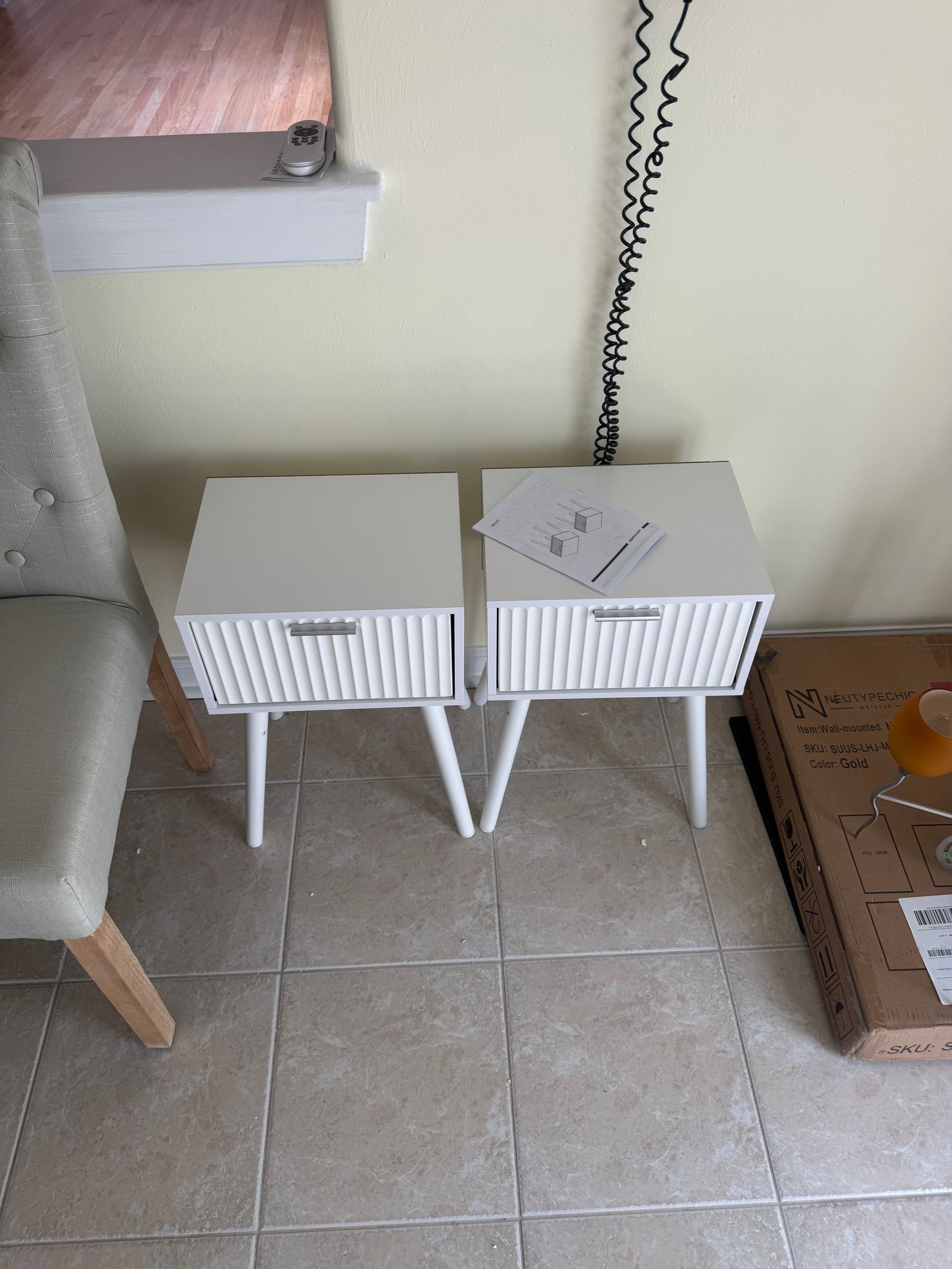 Two small white storage boxes on legs beside a wall outlet and cardboard box on a tiled floor.