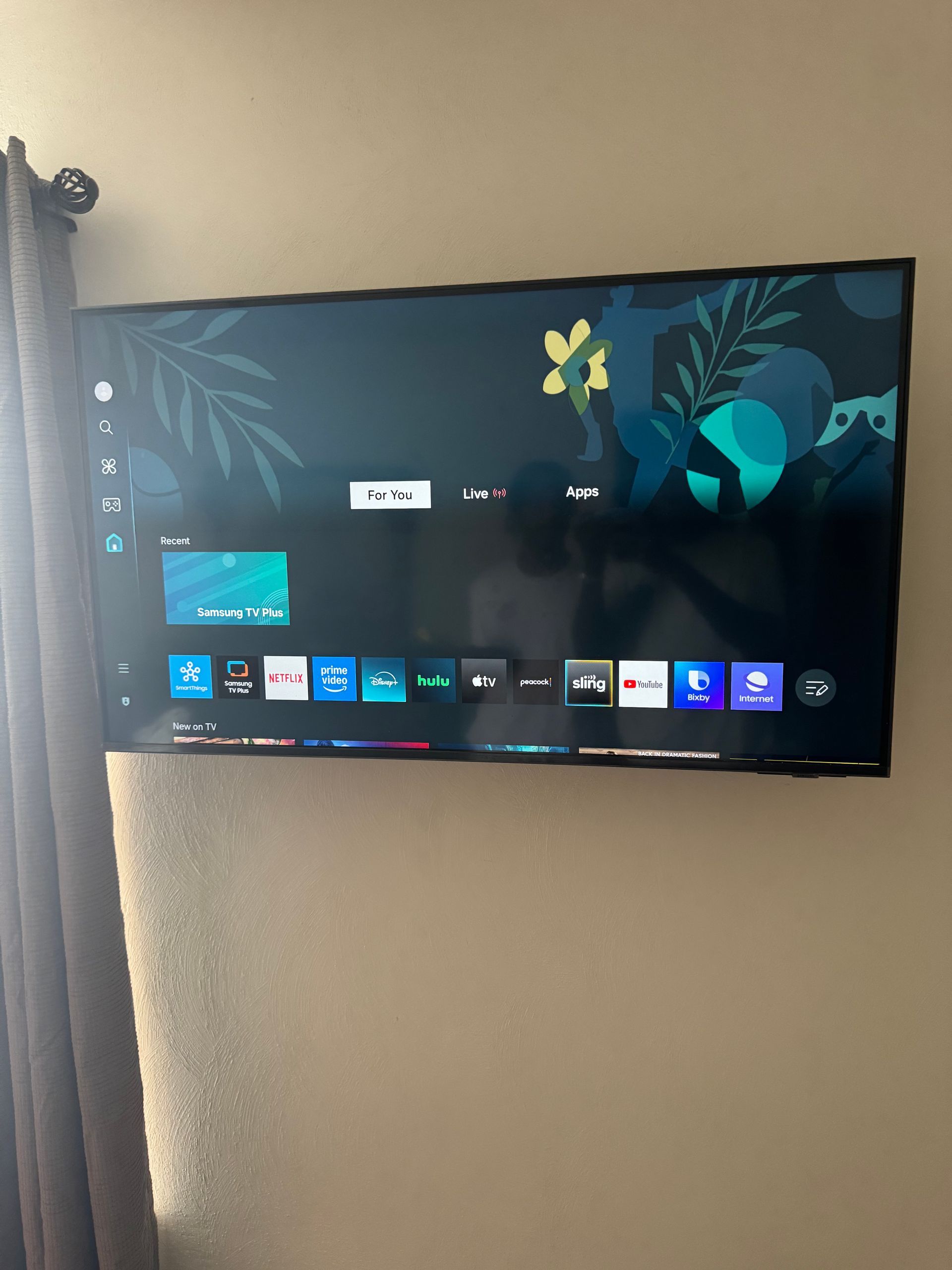 Wall-mounted TV showing a home screen with app icons on a beige wall.
