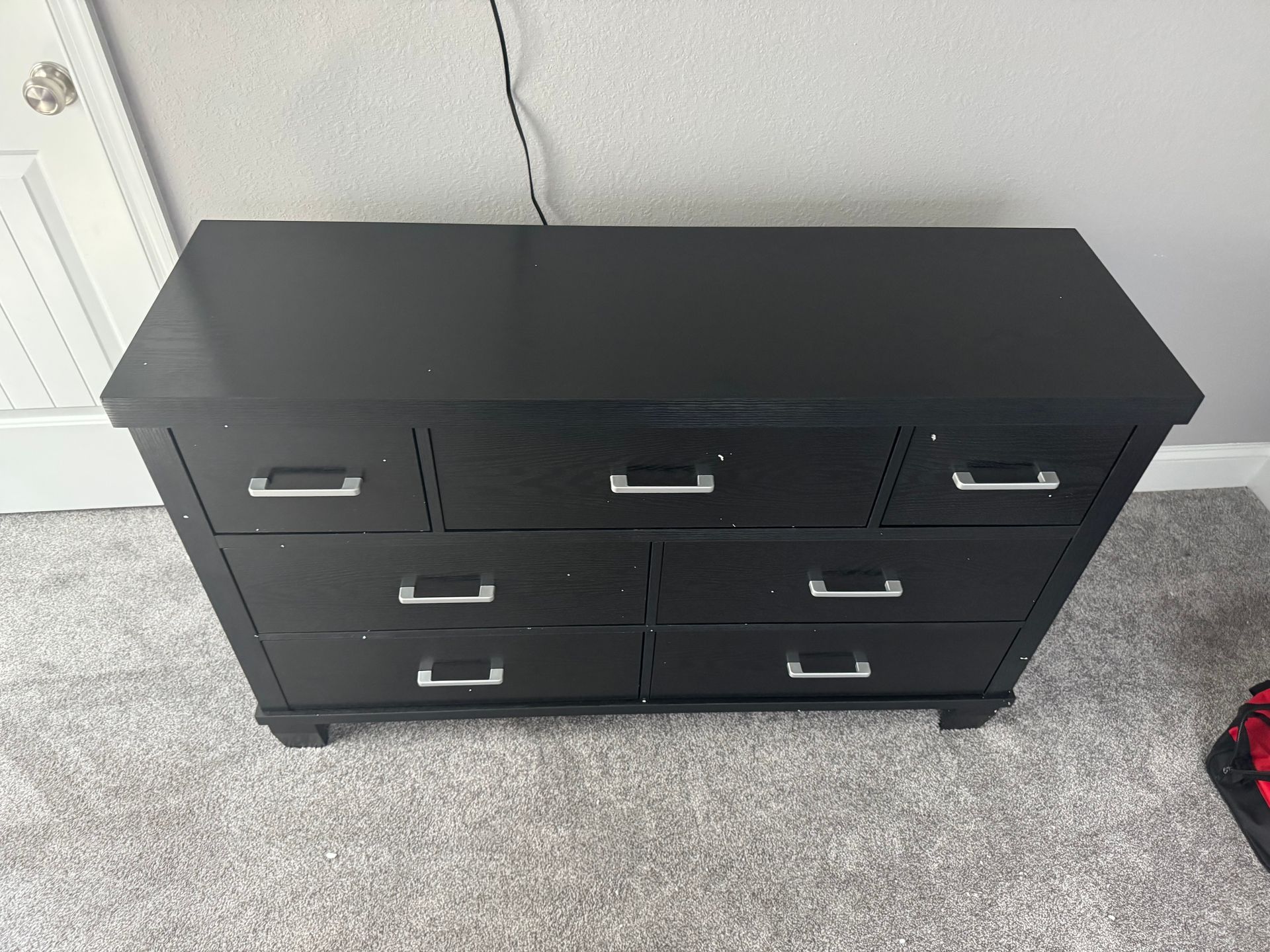 Black six-drawer dresser against a gray wall on beige carpet, with a power cord above it