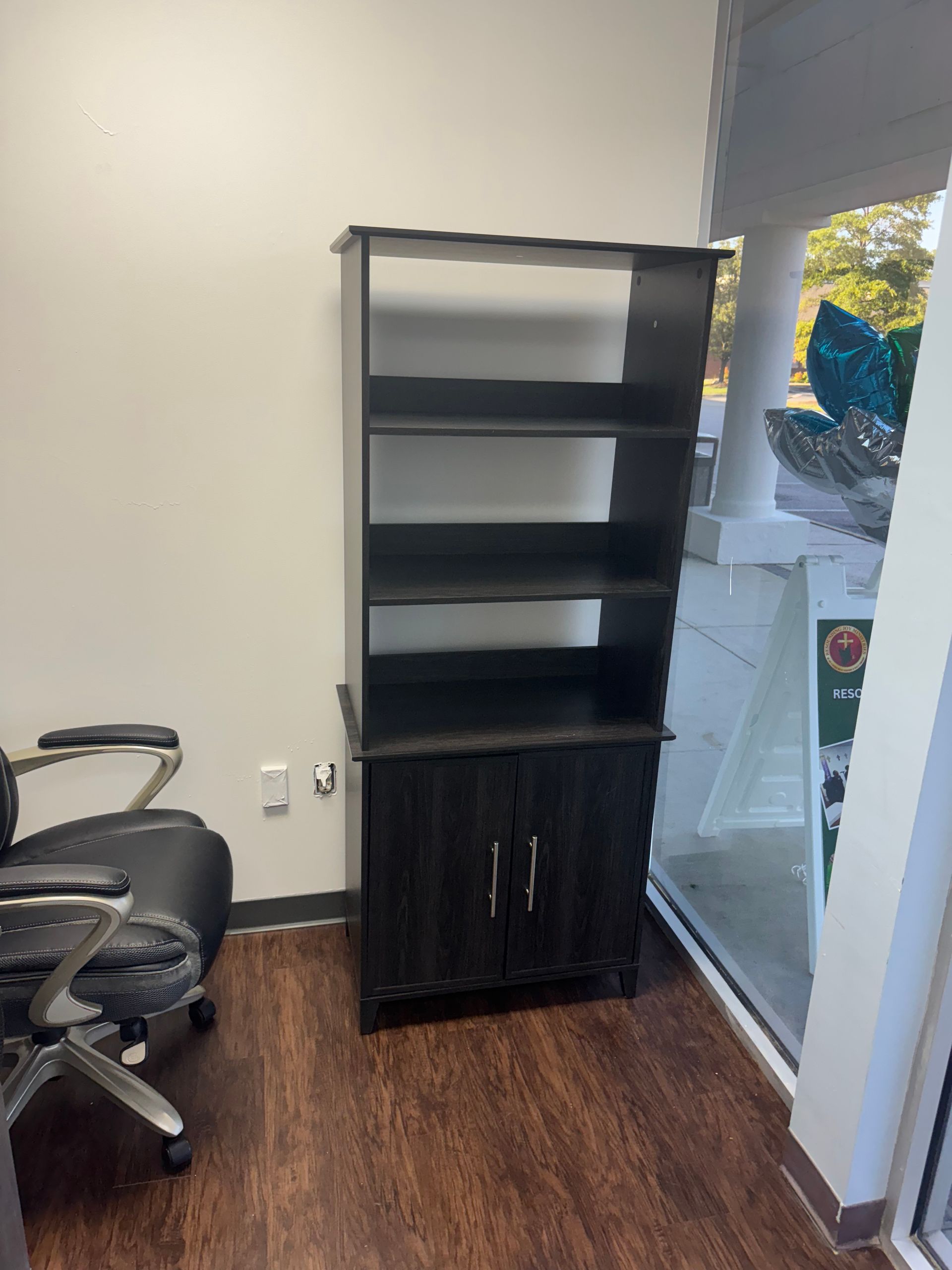 Black shelving unit with cabinet in an office corner beside a desk and glass door.