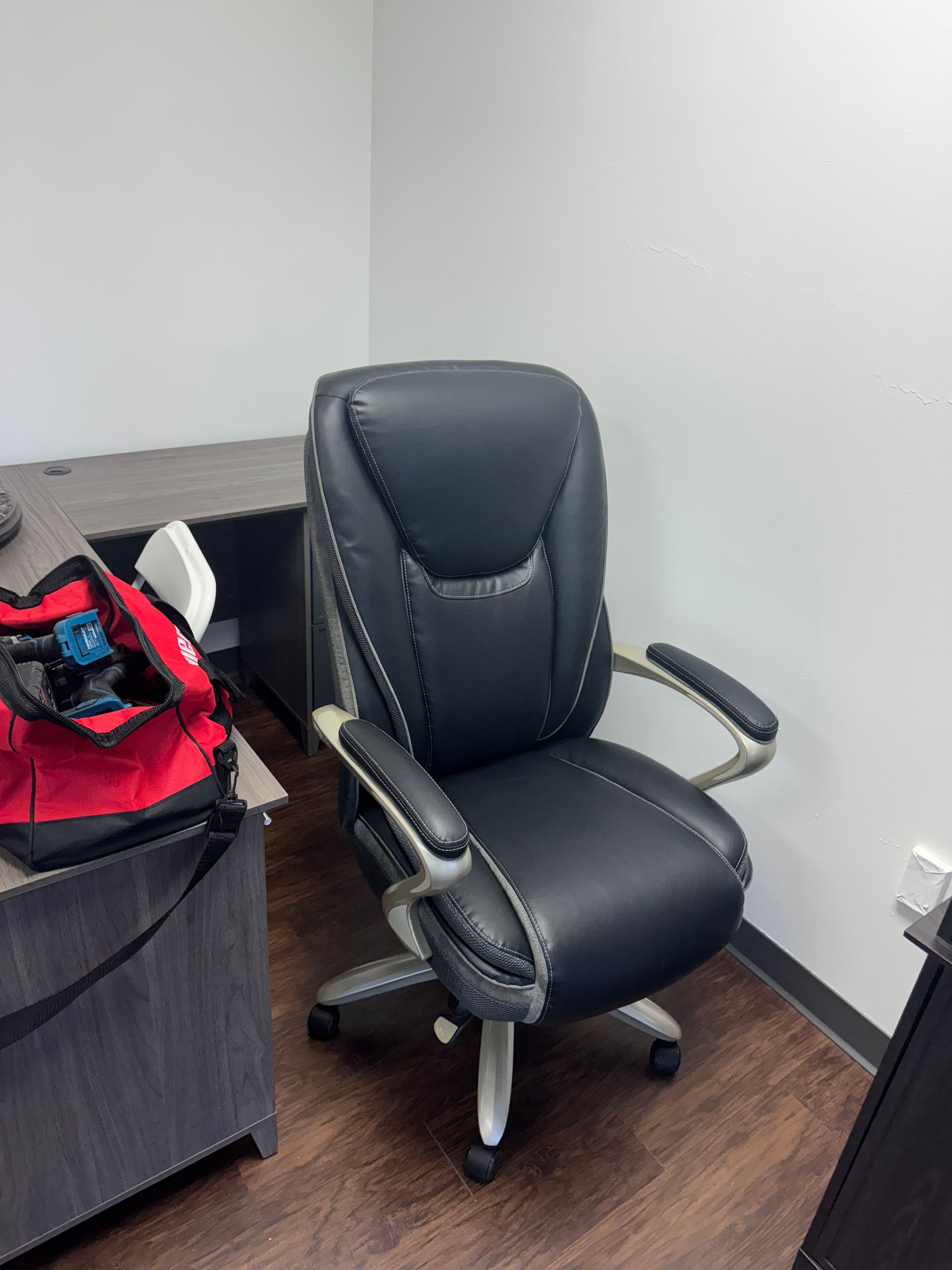 Black office chair beside a desk in a corner, with a red bag on the left.