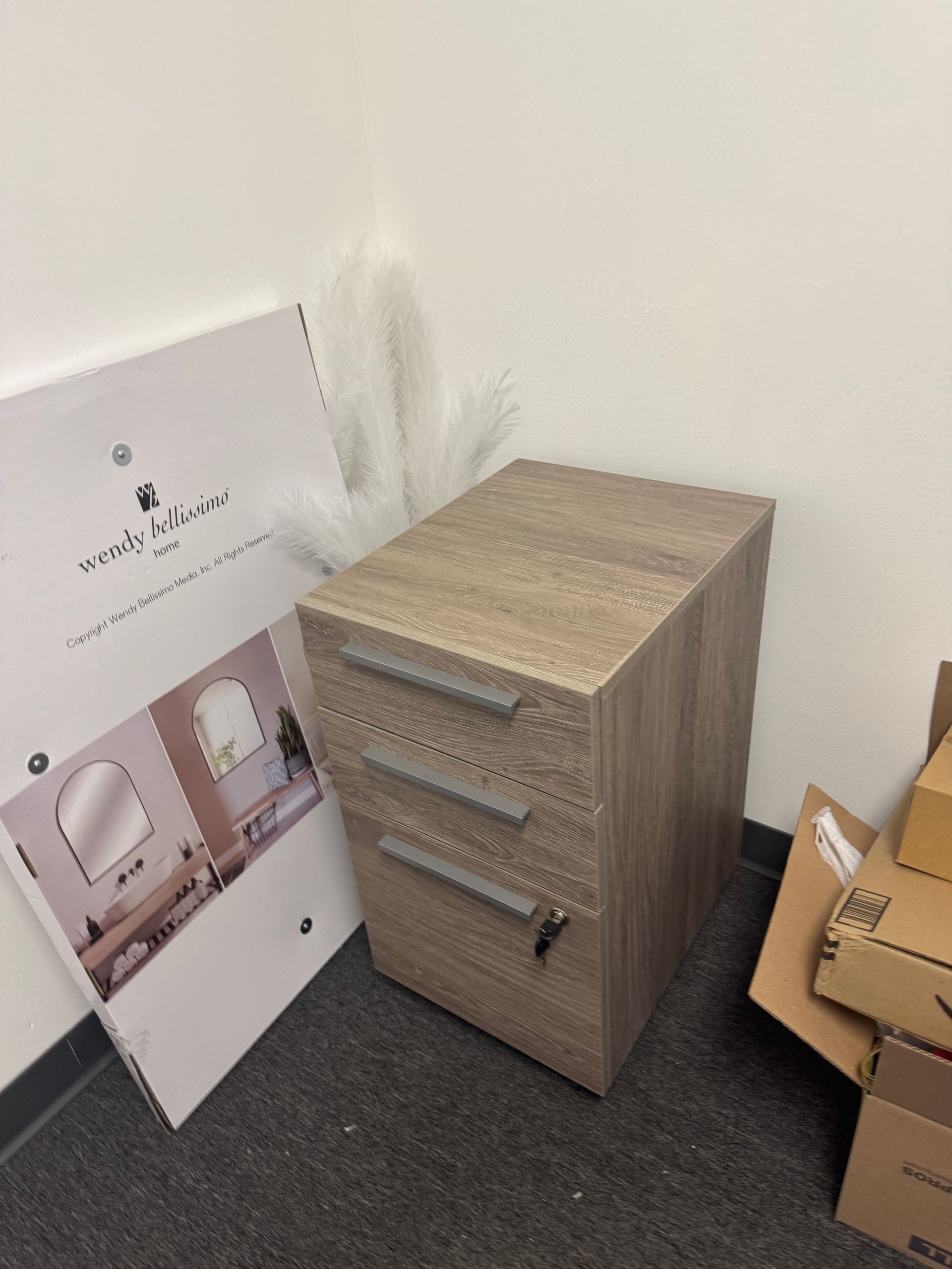 Beige three-drawer filing cabinet in a corner, with a white board leaning beside it and cardboard boxes nearby.
