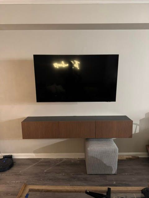 Wall-mounted TV above a floating wooden console with a gray ottoman in a minimalist room.