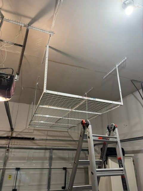 Ceiling-mounted wire storage rack being installed in a garage, with a ladder underneath.