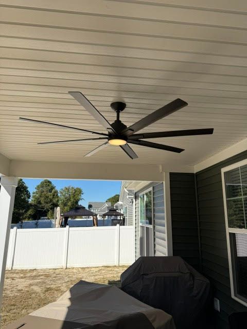 Covered patio with a ceiling fan, grill, white fence, and yard in the background.