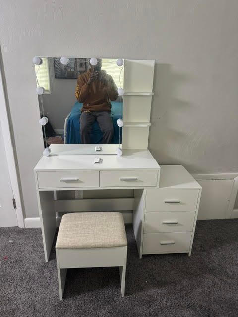 White vanity table with mirror, drawers, stool, and chair in a room; a person sits in the reflection.