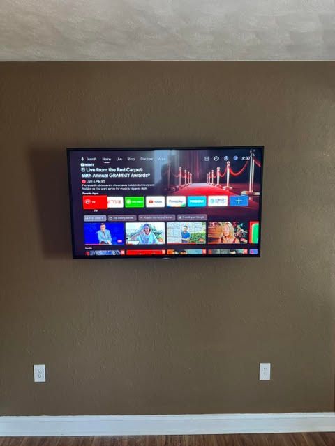 Wall-mounted TV displaying a streaming home screen on a beige wall with white baseboards.
