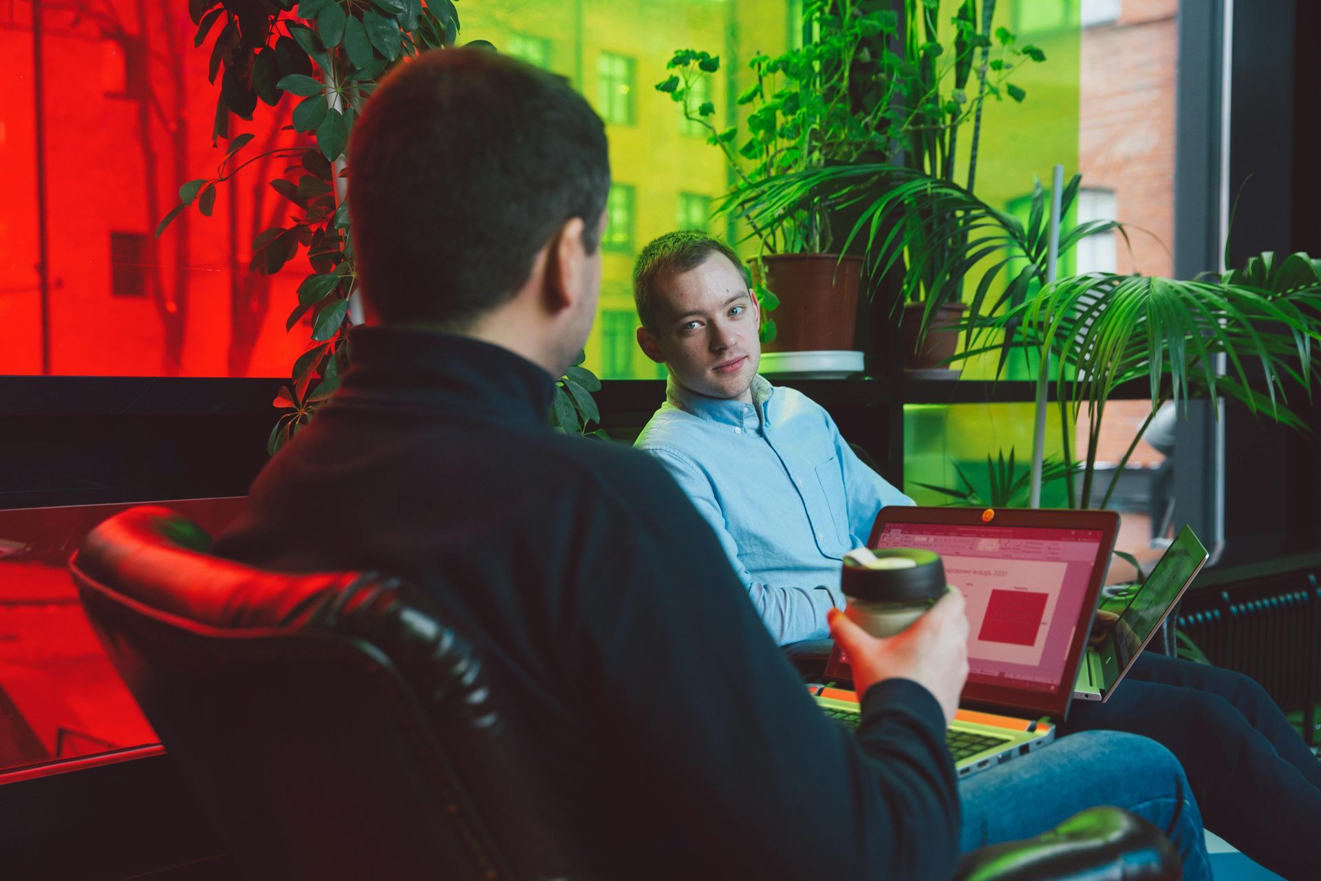 Two men at a table with laptops, talking. One holds a coffee cup. Green plants, colorful window in background.