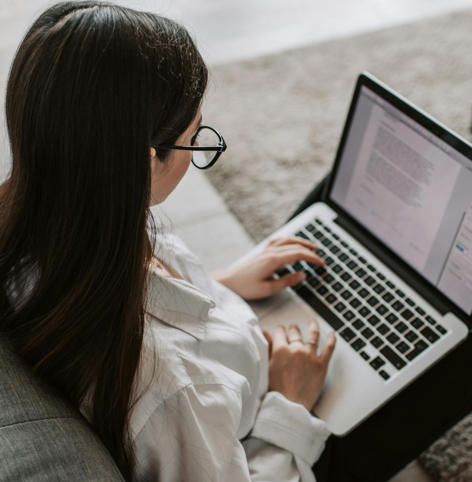 Woman with glasses typing on a laptop, sitting on a couch.