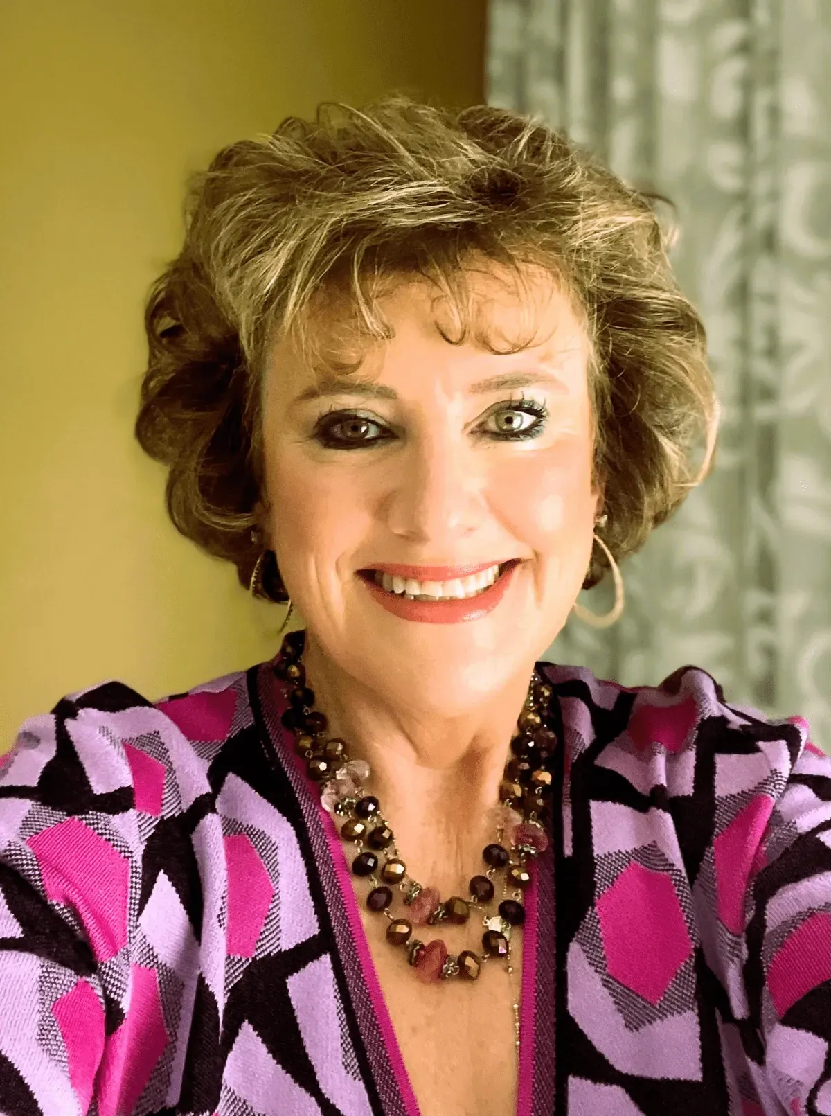 Woman with short, curly hair smiles, wearing a pink patterned top and layered beaded necklace.