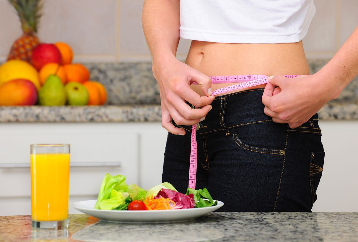 Woman measuring her waist with a pink tape measure, healthy food, and a juice glass in a kitchen setting.