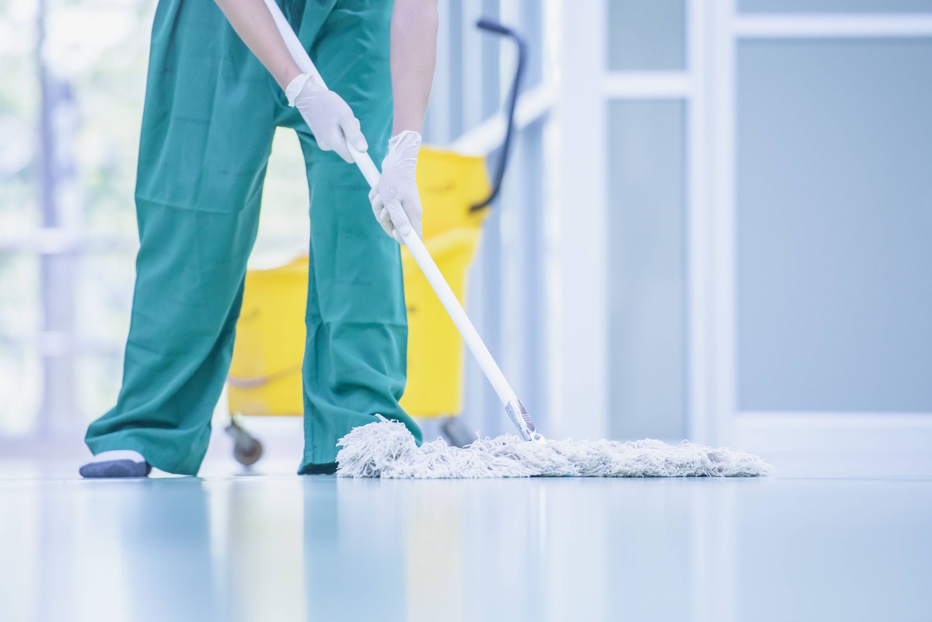 Person in green scrubs mops a shiny floor. A yellow bucket and wringer are in the background.