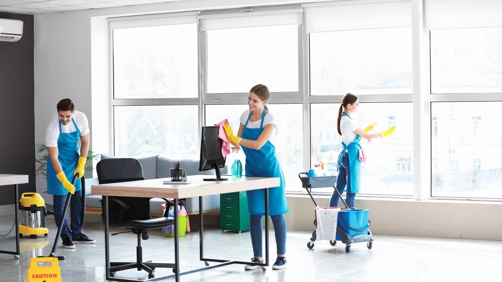 Three people cleaning an office; one vacuums, one dusts a desk, and one cleans a window.