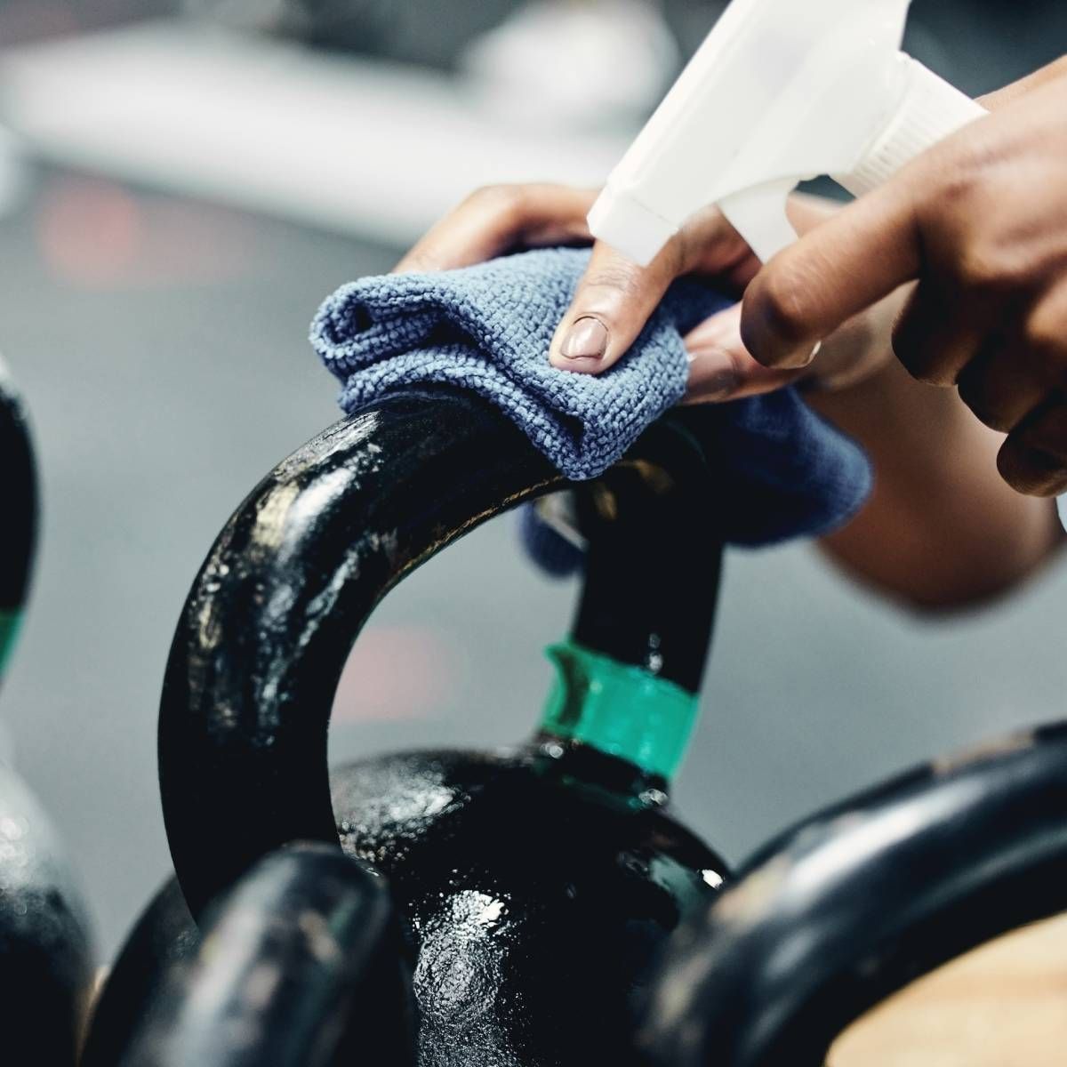 Person sanitizing a black kettlebell with a blue cloth and spray bottle in a gym.