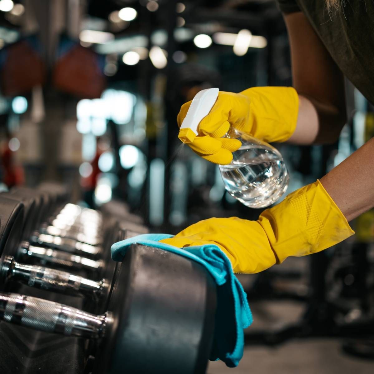 Person in yellow gloves sprays and wipes down a dumbbell in a gym.