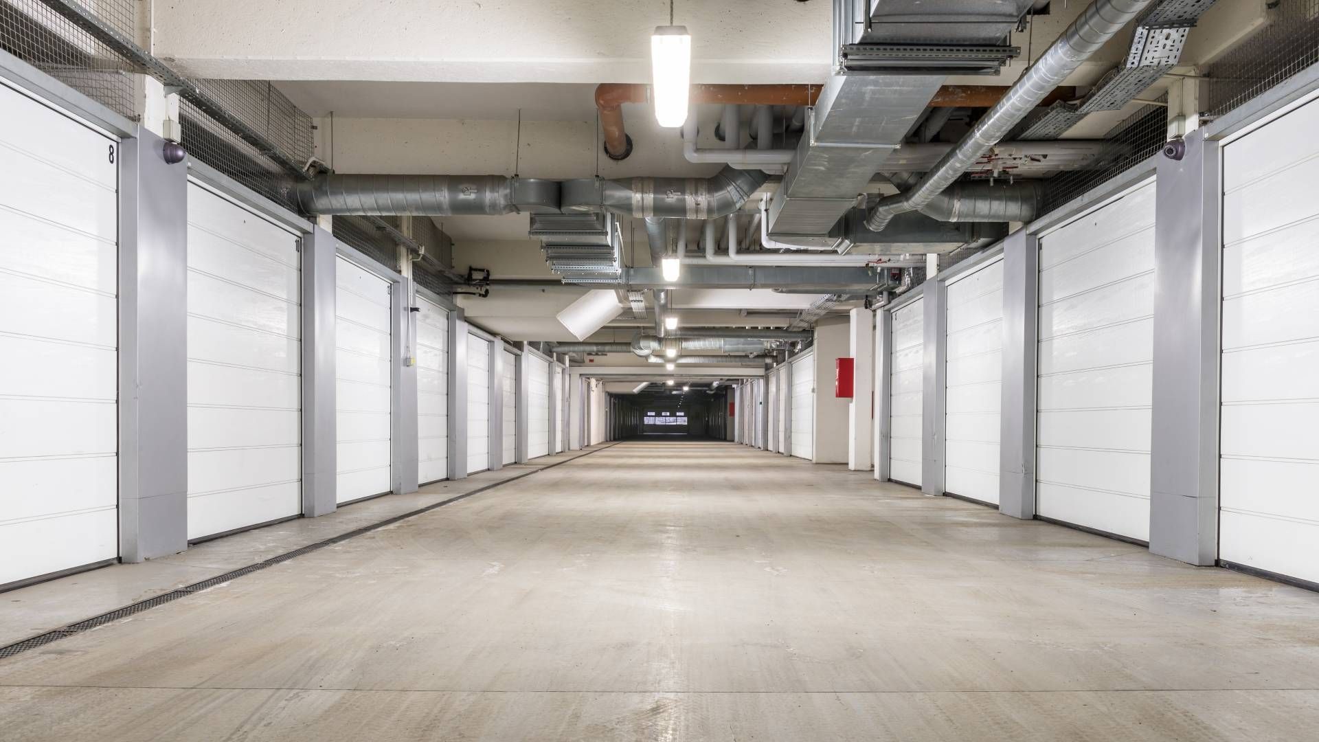 Long hallway of storage units with white doors, overhead lights, and exposed pipes.