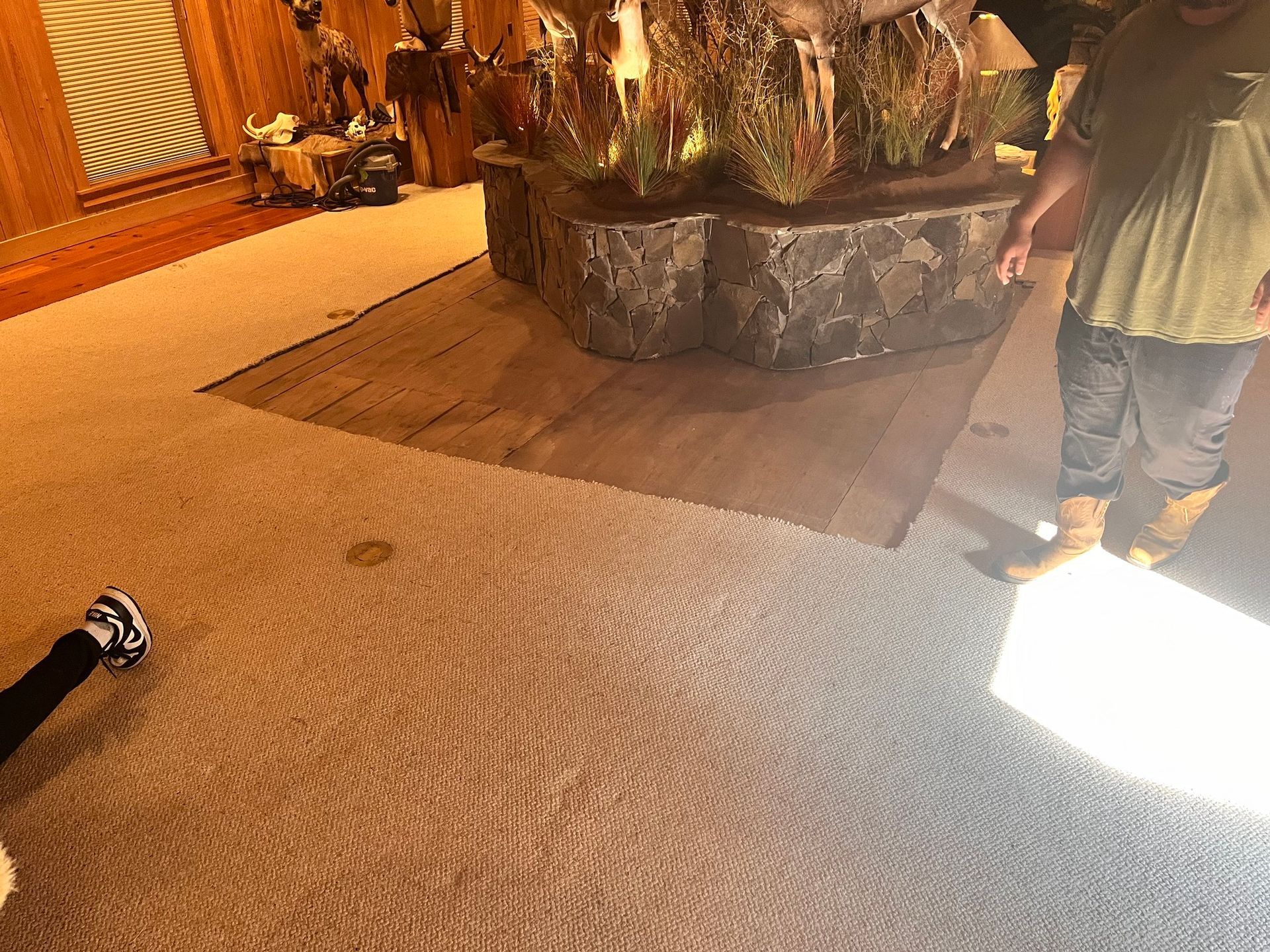 A person standing near a stone structure in a room with carpet and wood flooring, another person's legs are visible nearby.