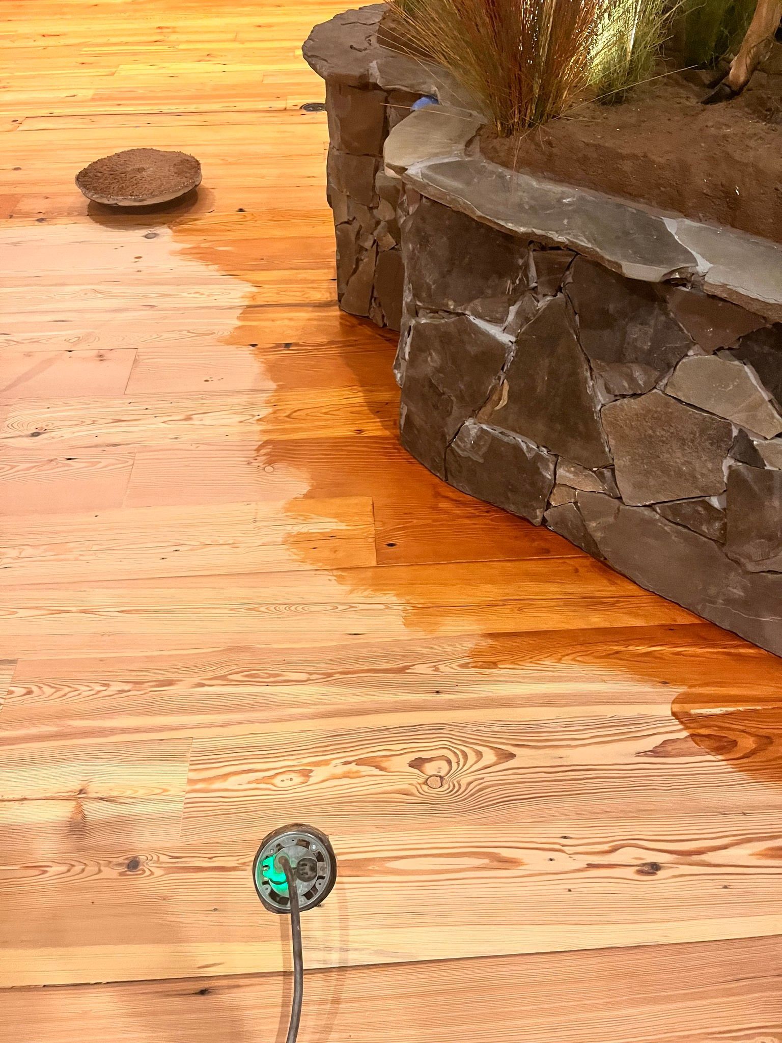 A wet wooden deck and stone planter box. A sprinkler waters the deck, leaving a sheen of water.