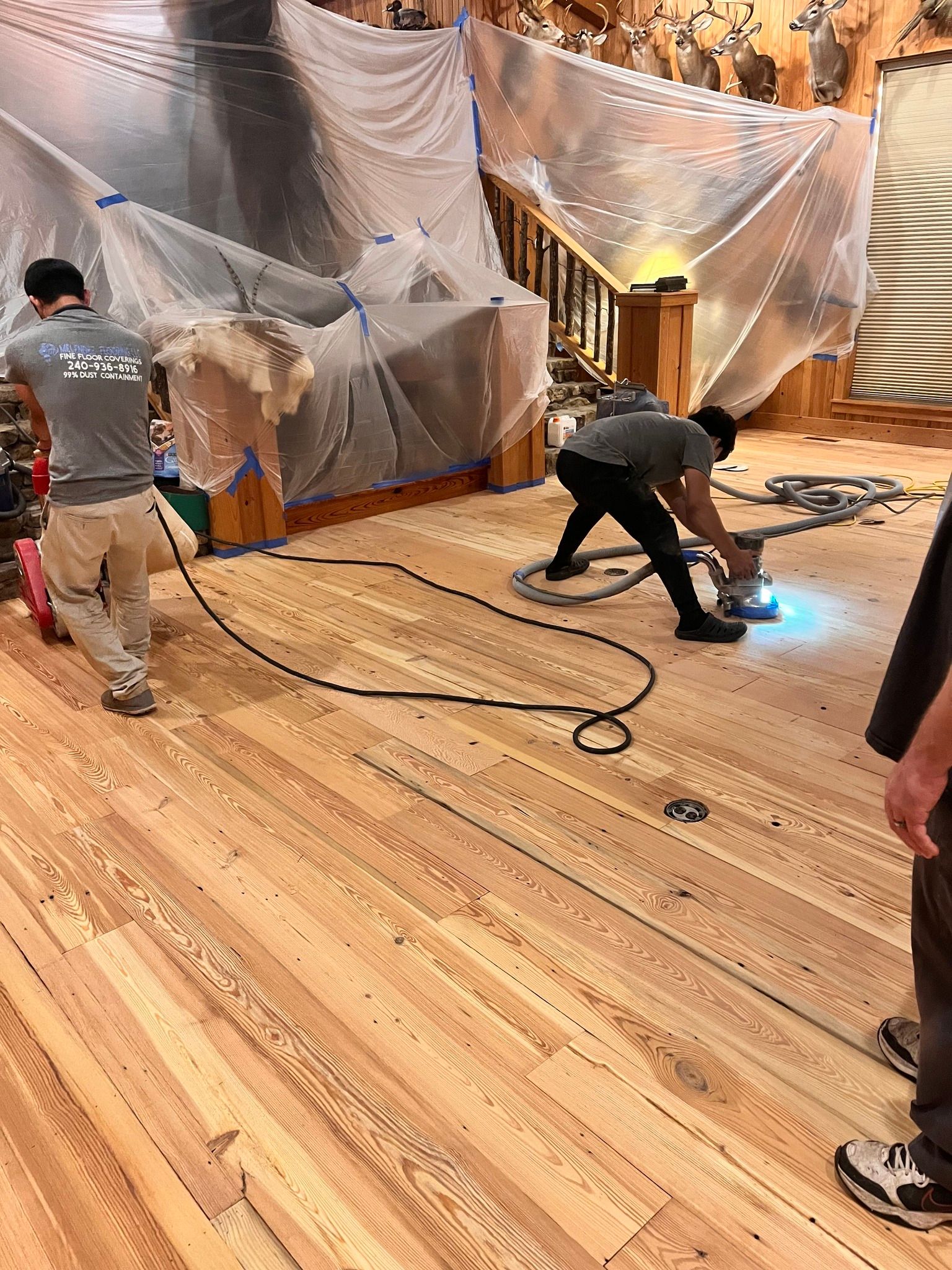 Workers sanding a wooden floor in a room. One operates a floor sander, while another tends to a power cord.