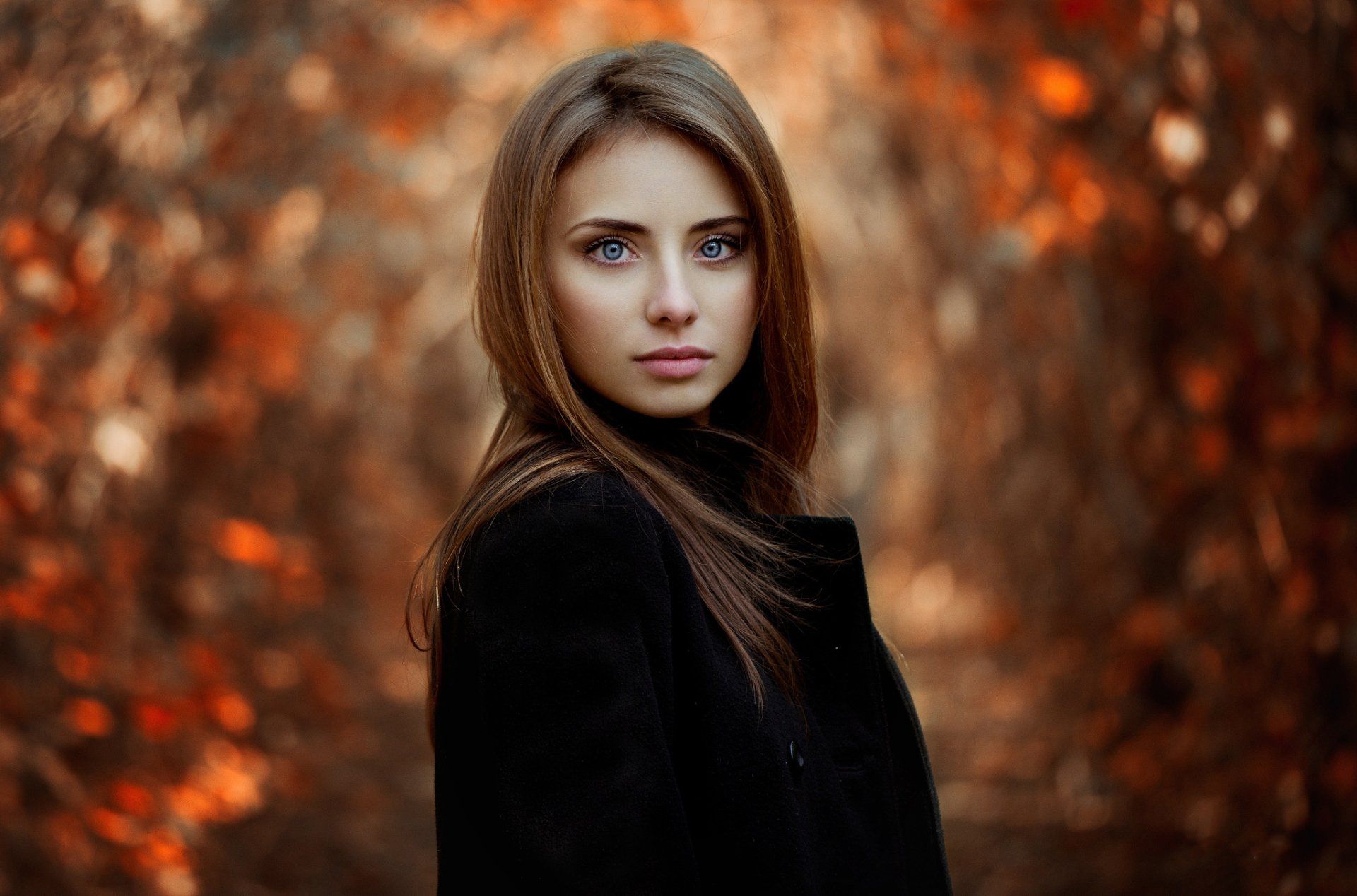 a woman in a black coat is standing in a forest looking at the camera .