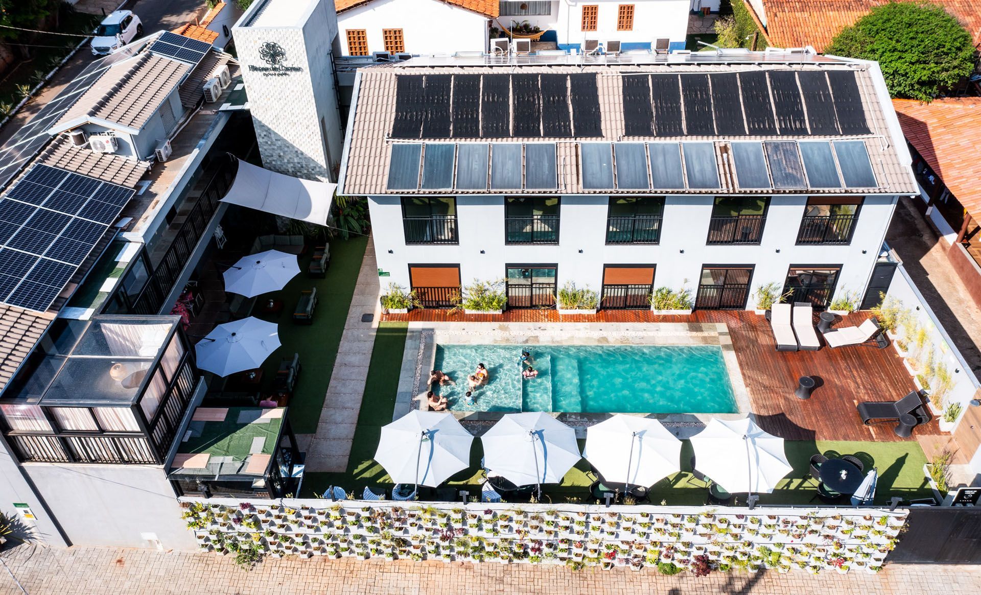 Vista aérea de um prédio branco de vários andares com piscina, painéis solares e guarda-sóis.