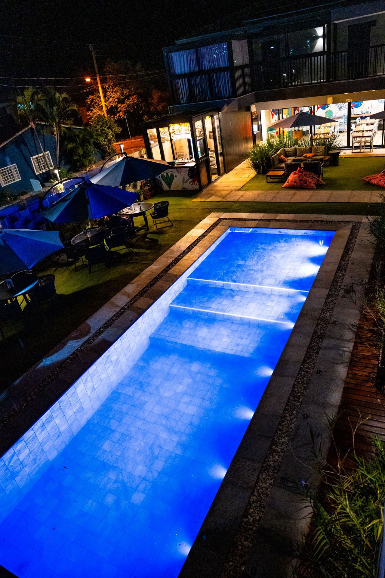 Vista noturna de uma piscina retangular iluminada com luzes azuis, perto de um prédio de dois andares e uma área de estar externa.