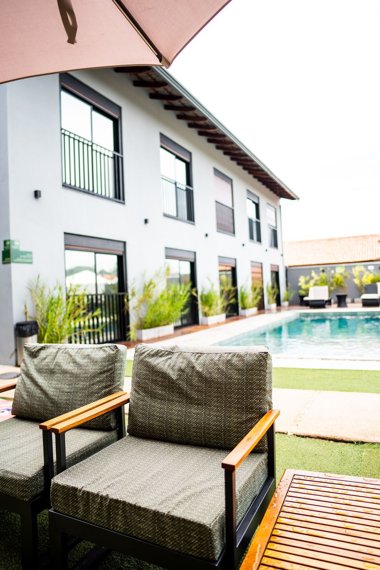 Duas cadeiras de vime e uma mesa de madeira ao lado de uma piscina, com um prédio de cor clara ao fundo.