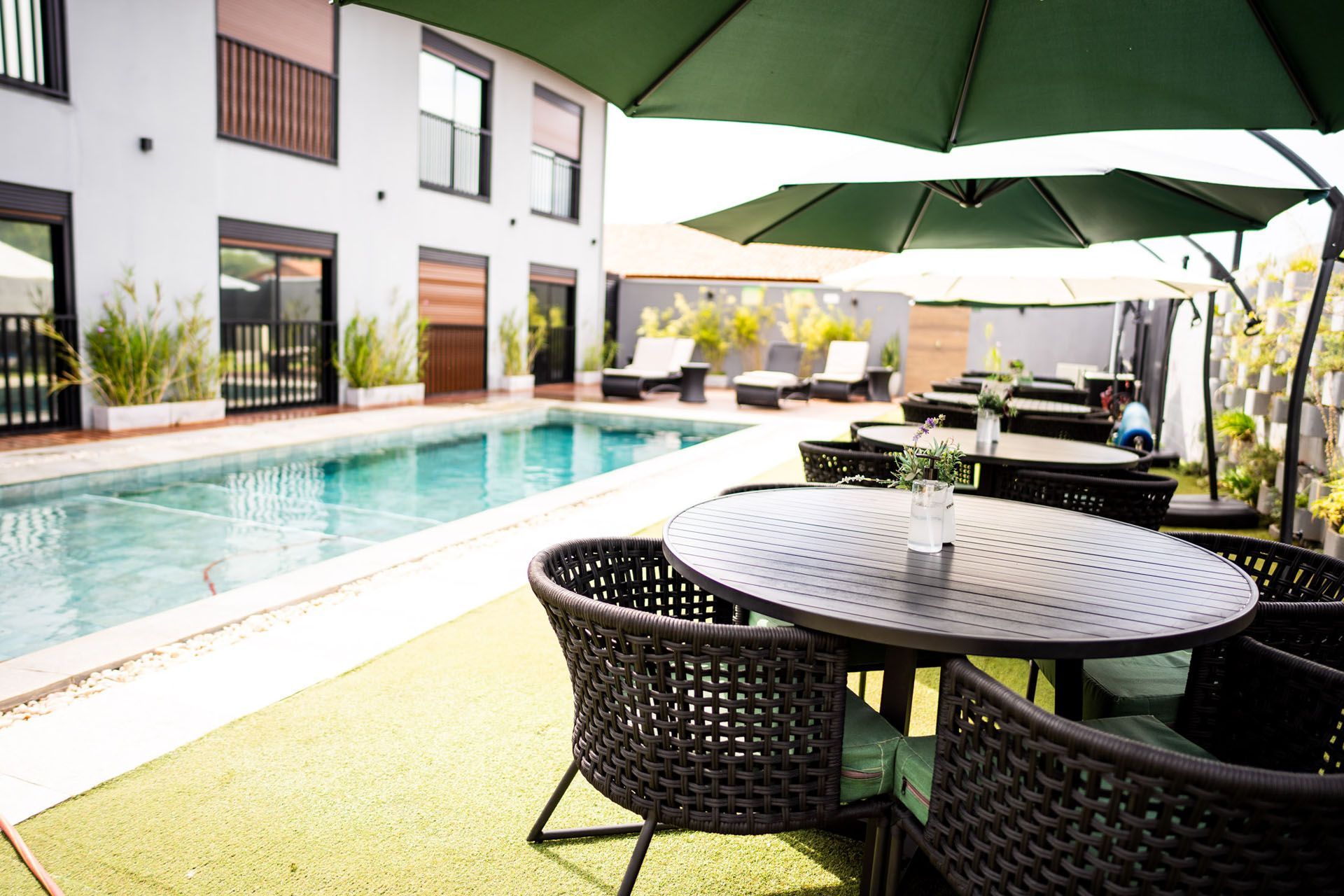 Área da piscina com mesa e cadeiras sob guarda-sóis; prédio ao fundo.