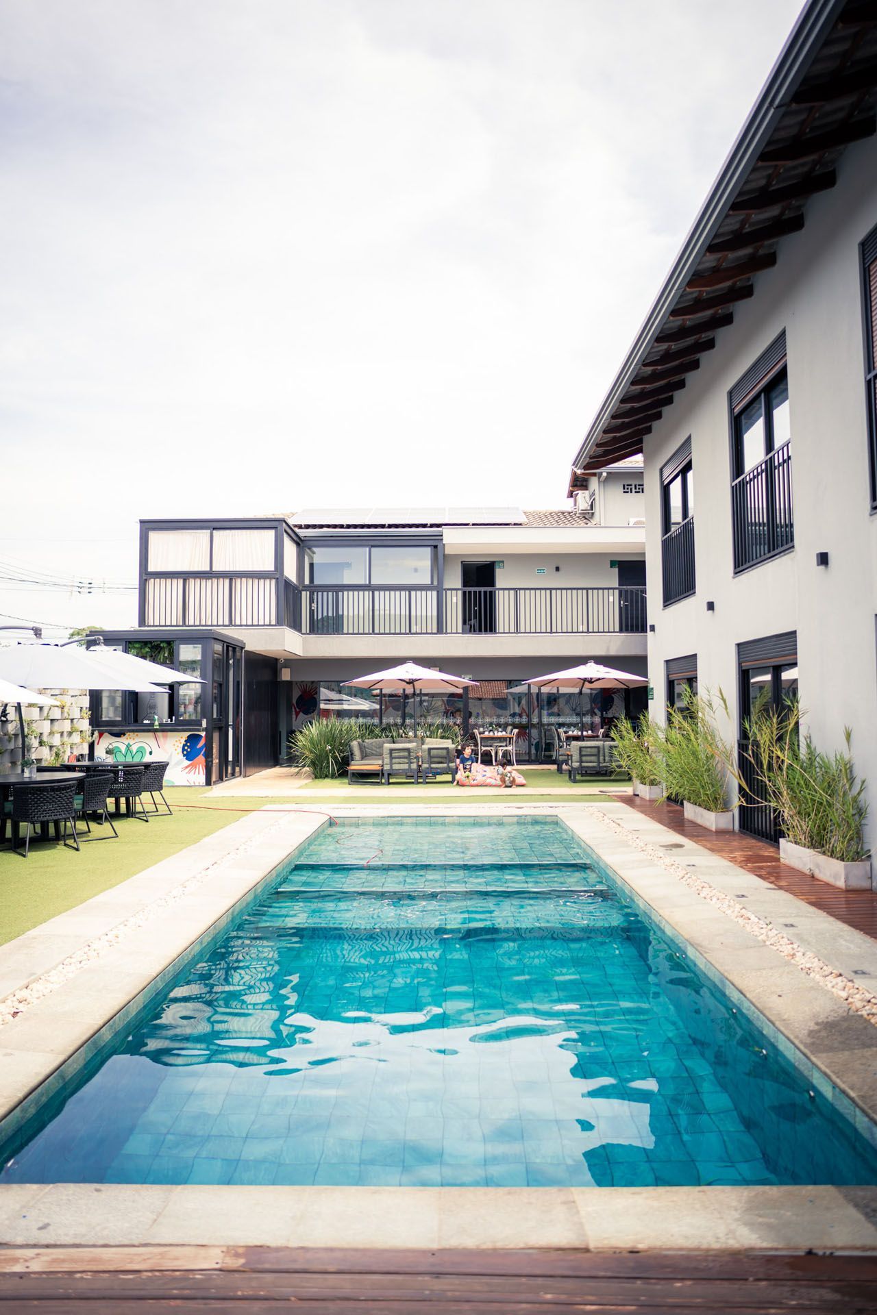 Piscina em frente a um prédio branco com mesas e cadeiras ao ar livre, sob um céu nublado.