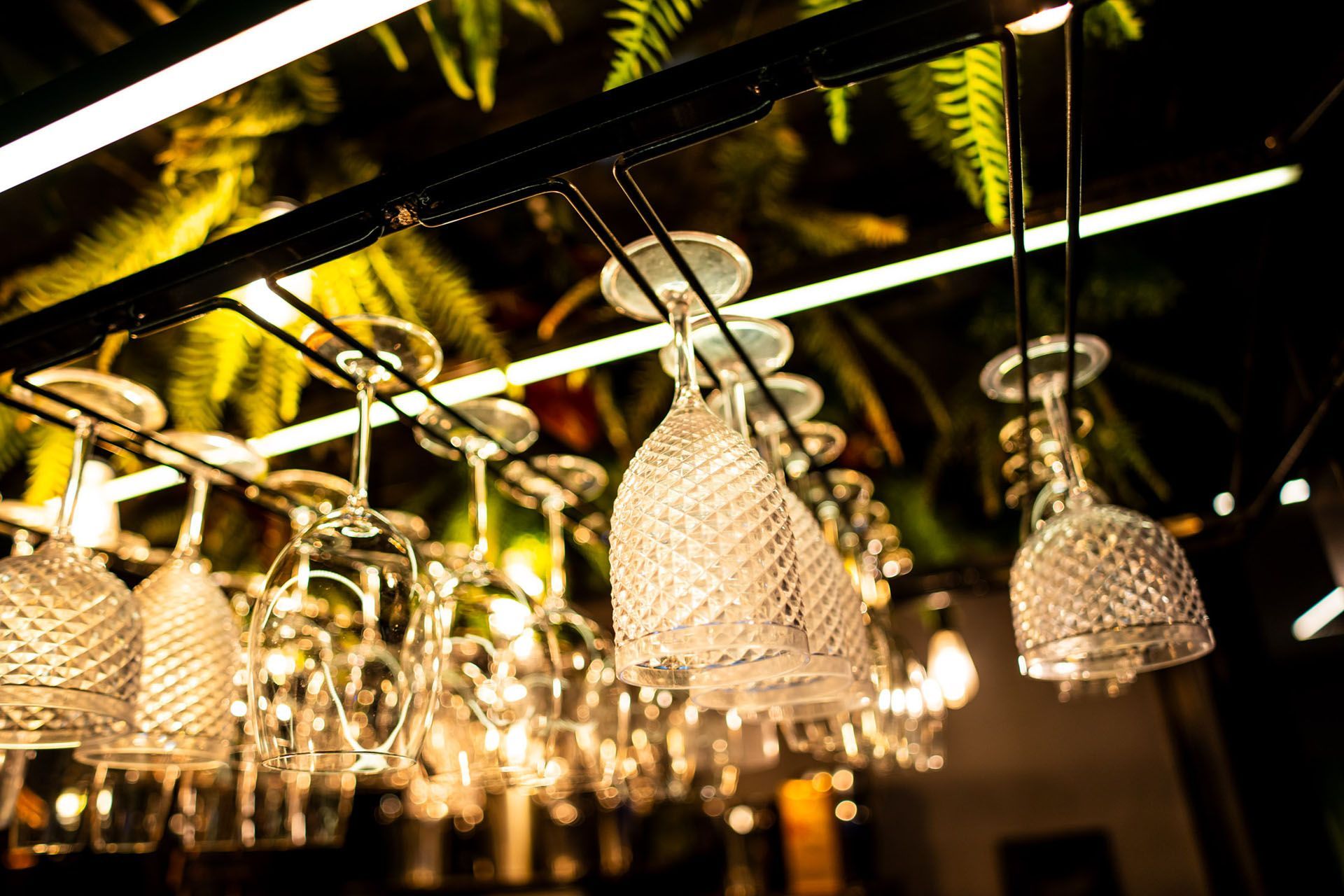 Taças de vinho penduradas de cabeça para baixo em um suporte, com um fundo desfocado de vegetação e luzes.