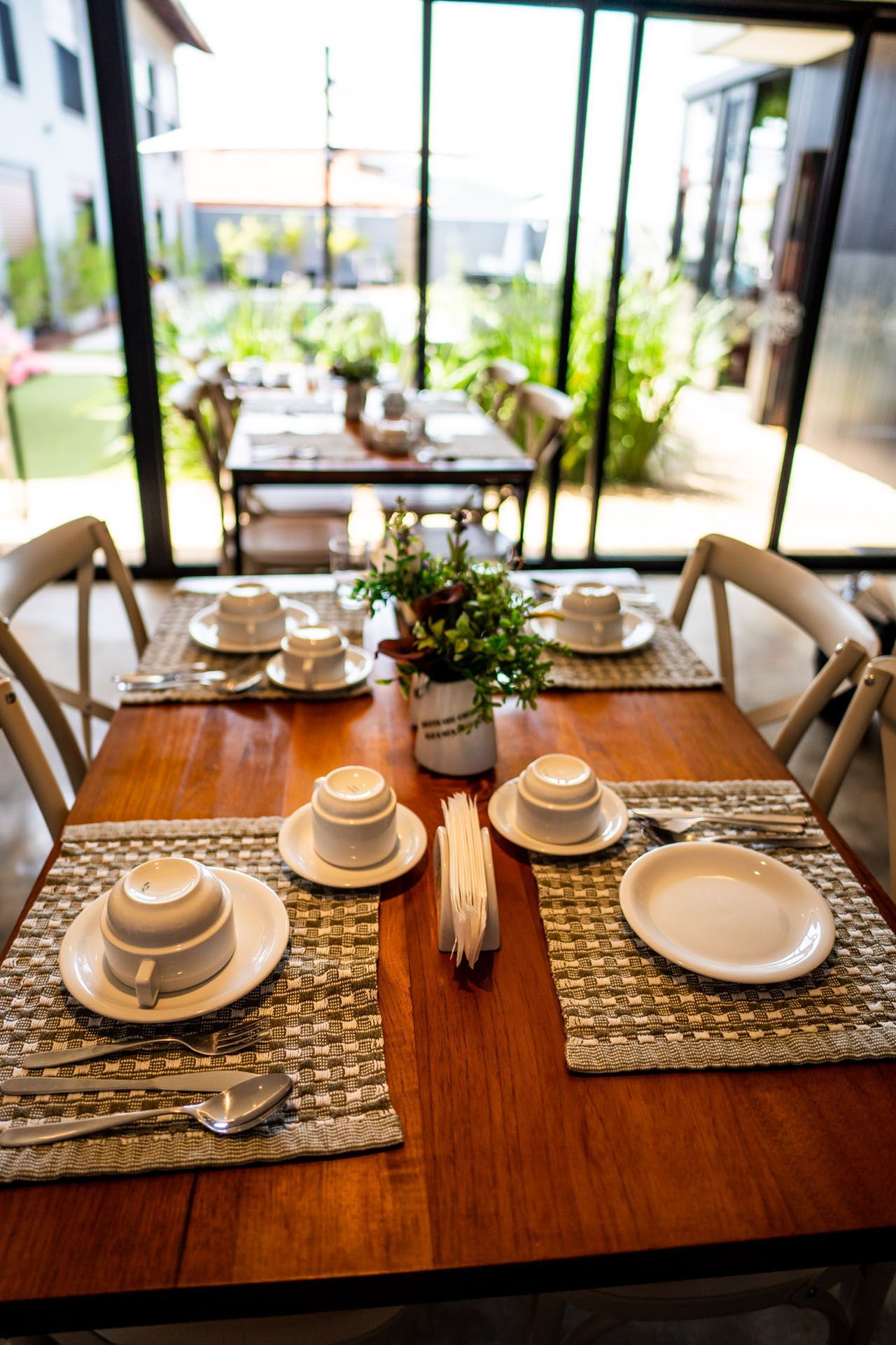 Mesa de madeira posta para refeição com pratos, xícaras e talheres em uma sala iluminada com vista.