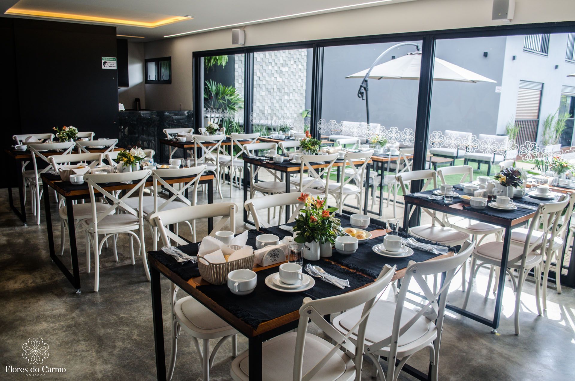 Sala de jantar do restaurante com mesas postas para o serviço, algumas com arranjos florais e cadeiras brancas.