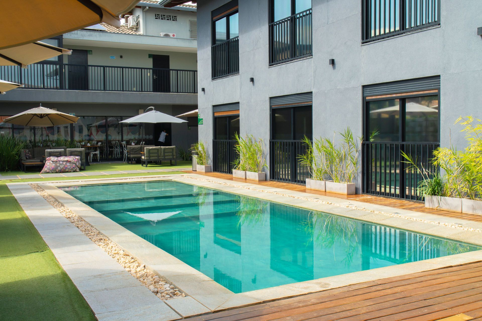 Piscina com água azul-turquesa rodeada por deck de madeira e gramado verde, ao lado de um prédio cinza.