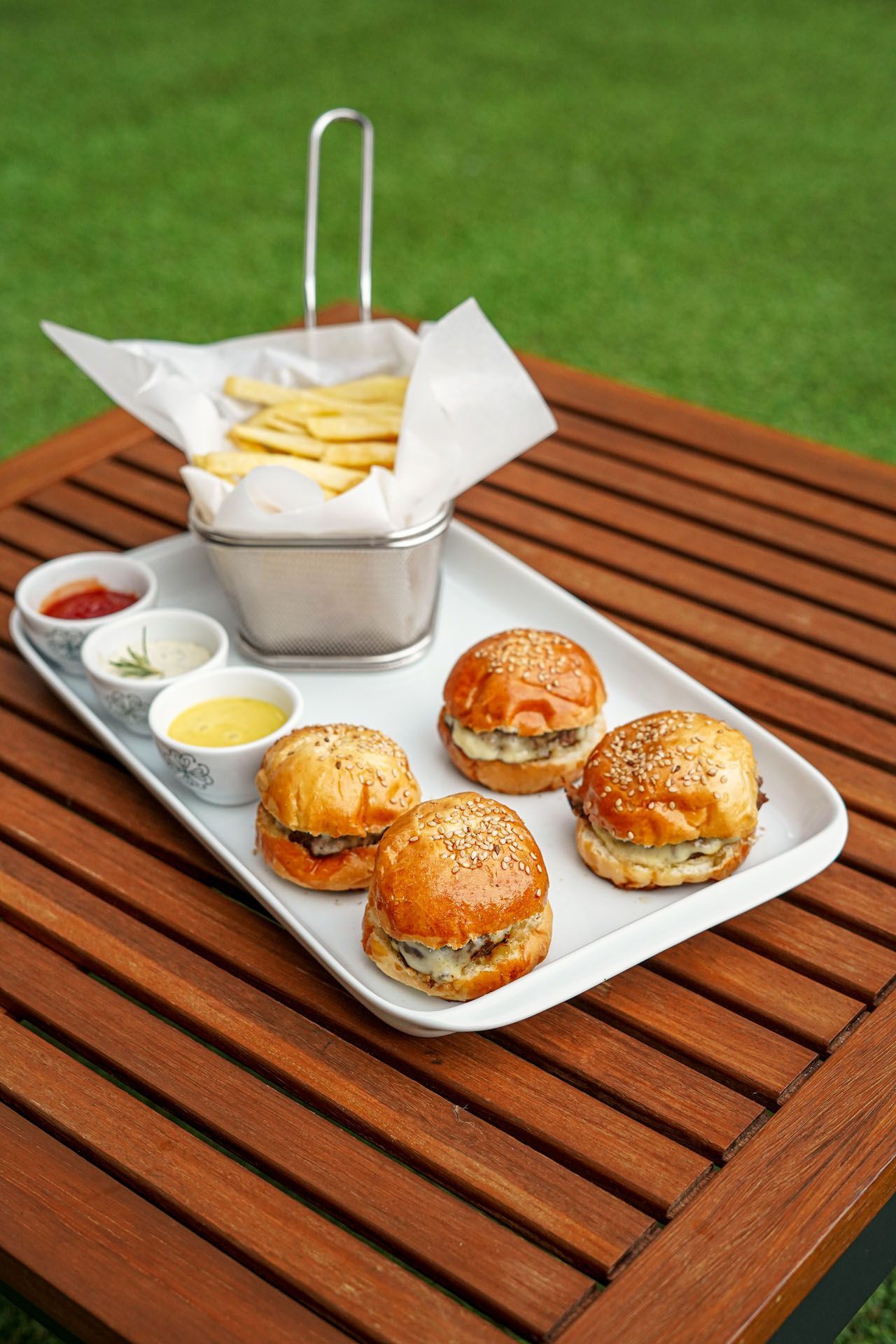 Mini hambúrgueres, batatas fritas e molhos para mergulhar sobre uma mesa de madeira, com grama ao fundo.