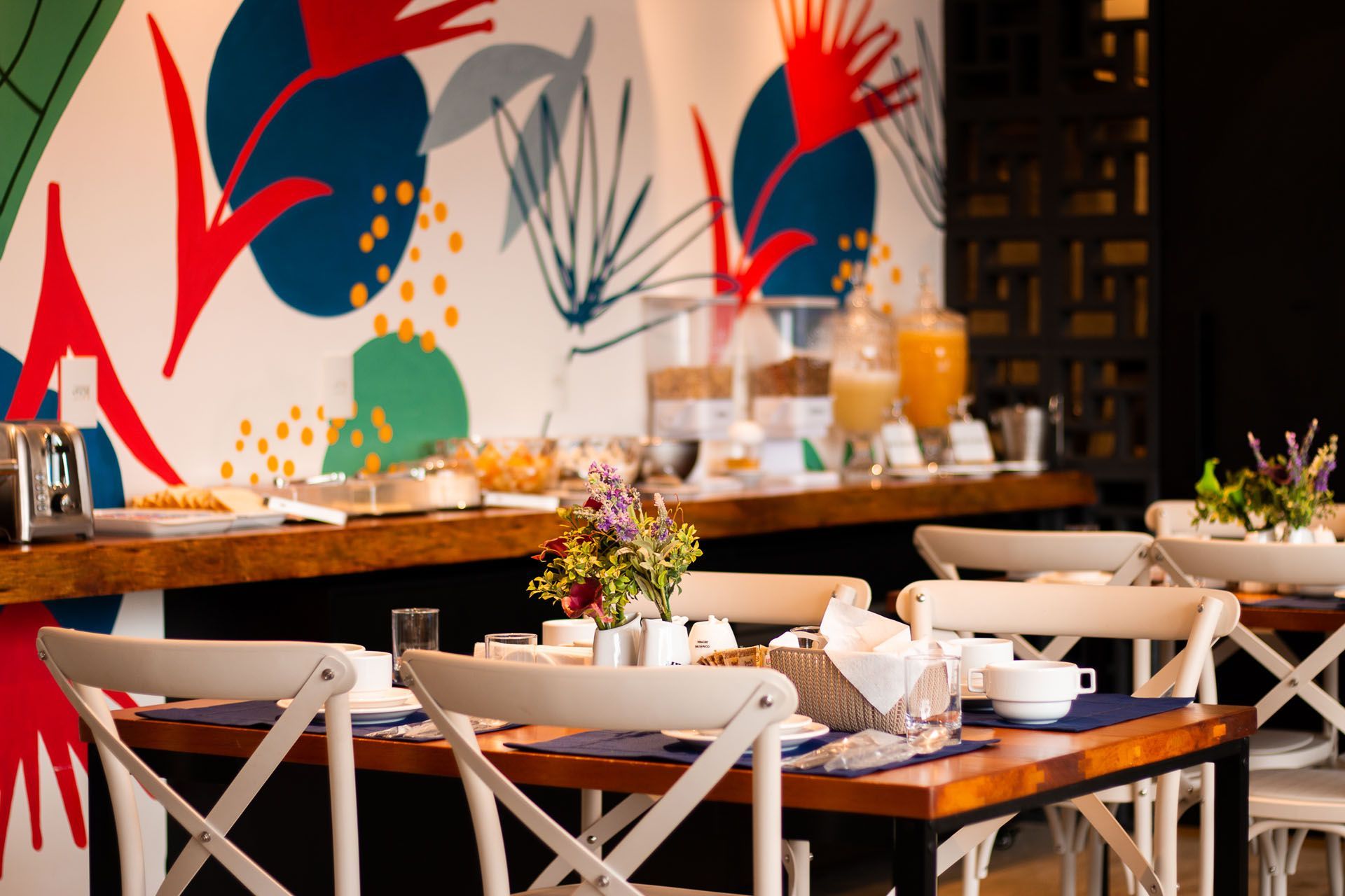 Buffet de café da manhã com mesas e cadeiras dispostas em frente a um mural colorido na parede.