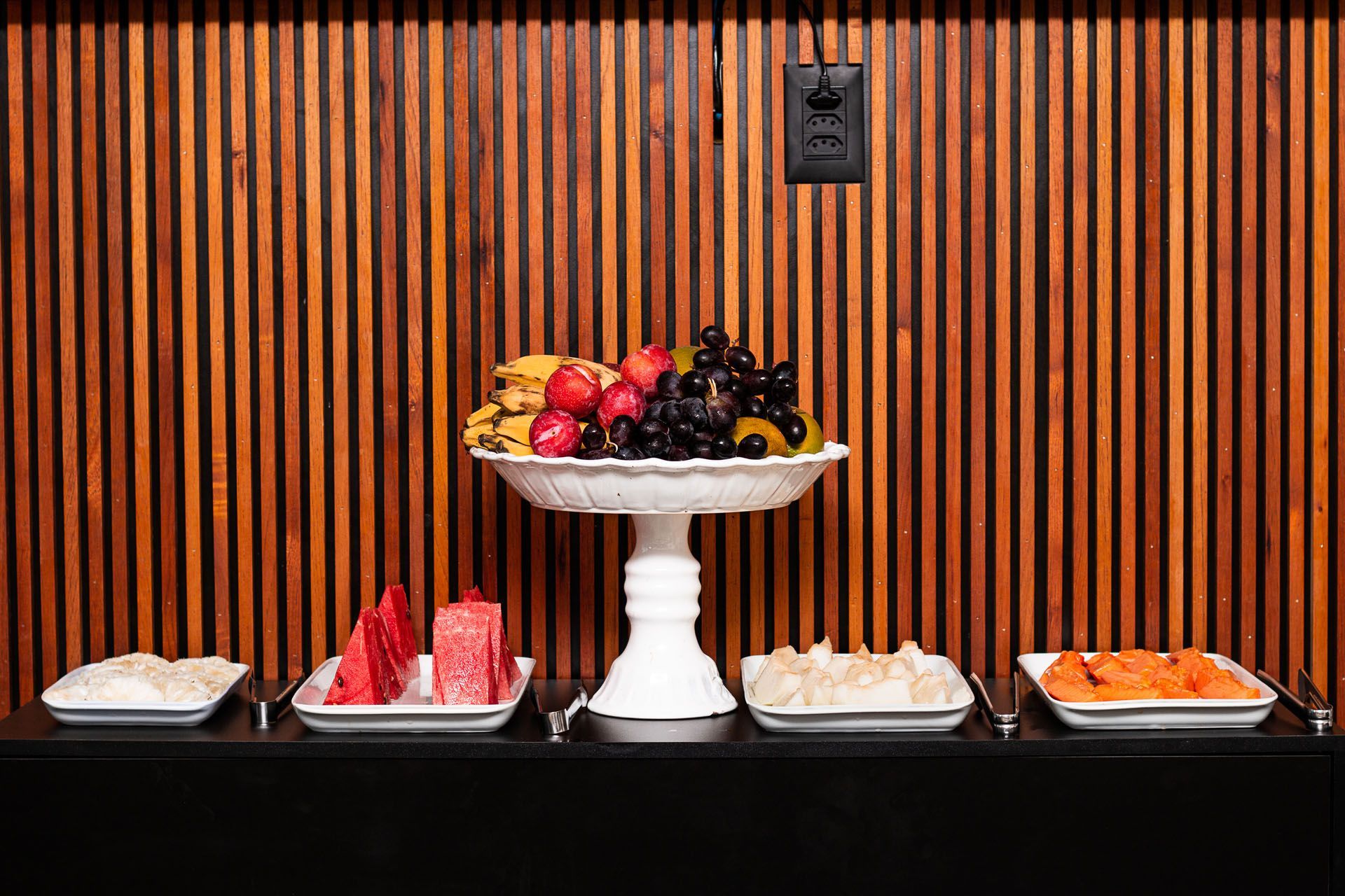 Bandeja de frutas com diversas frutas e pequenas tigelas brancas com alimentos sobre uma bancada preta, com painel de madeira ao fundo.