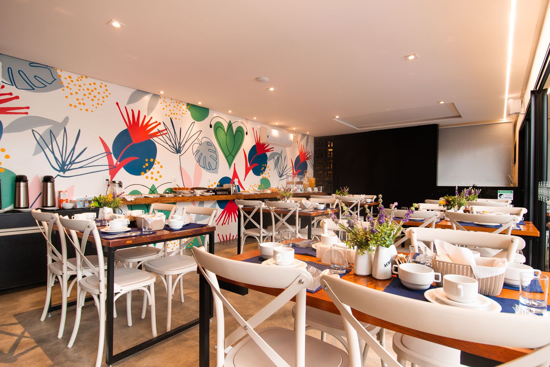 Sala de jantar com mesas de madeira, cadeiras brancas e um mural colorido. Aparelho de café da manhã à vista.