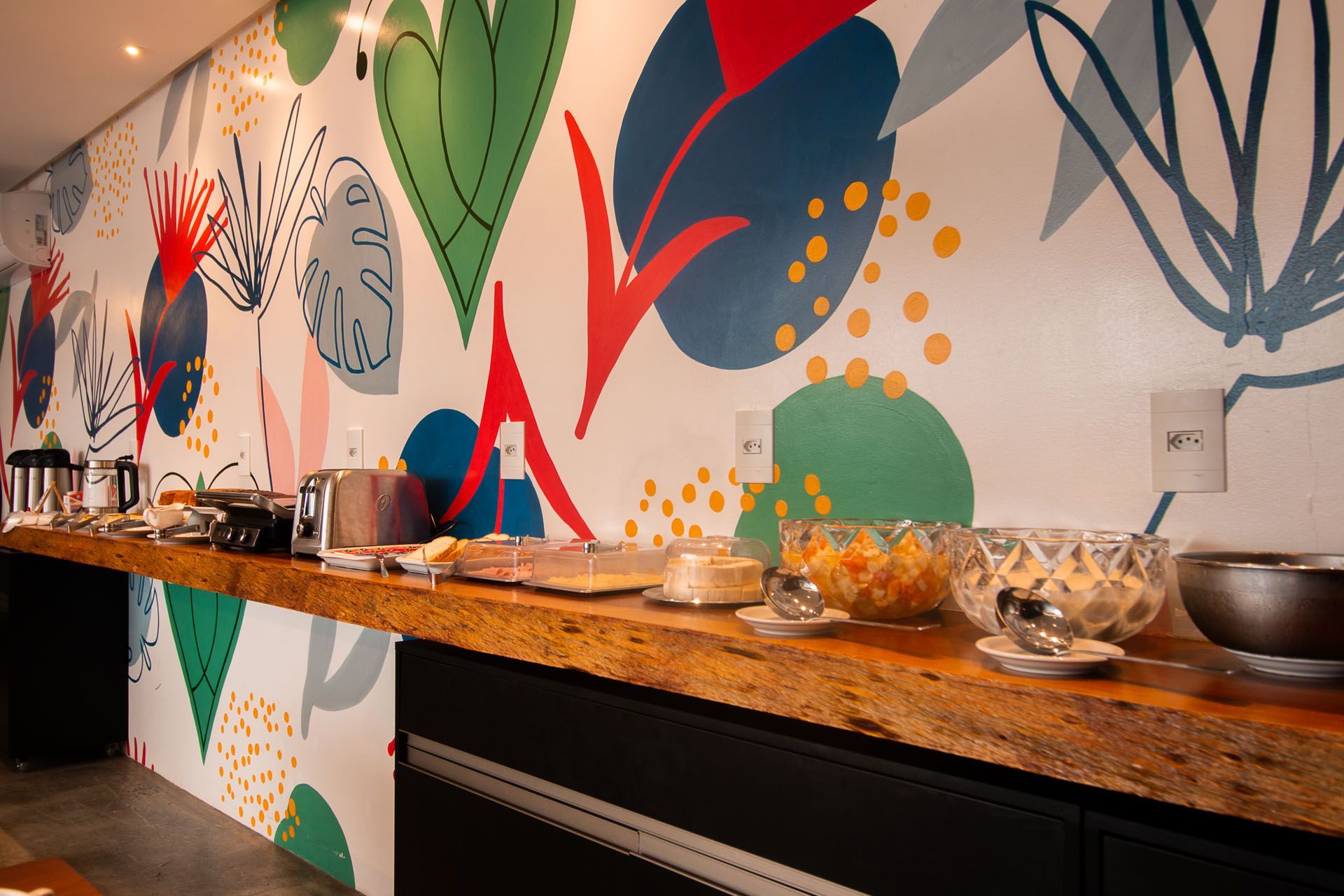 Buffet de café da manhã com diversas opções gastronômicas em um balcão de madeira, com um mural colorido ao fundo.