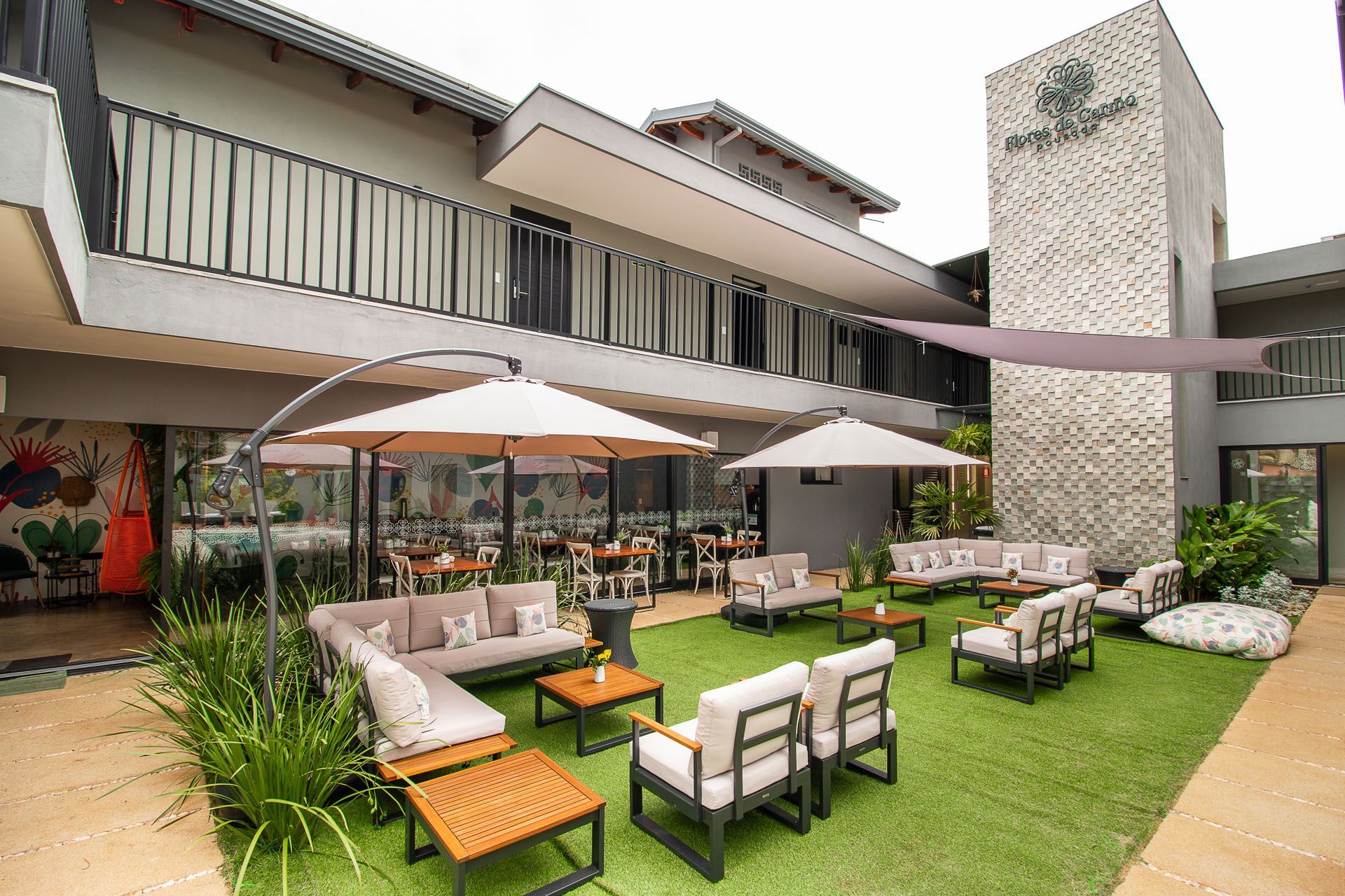 Área de estar externa com cadeiras, guarda-sóis e mesas de jantar perto de um prédio. Grama verde e um mural são visíveis.