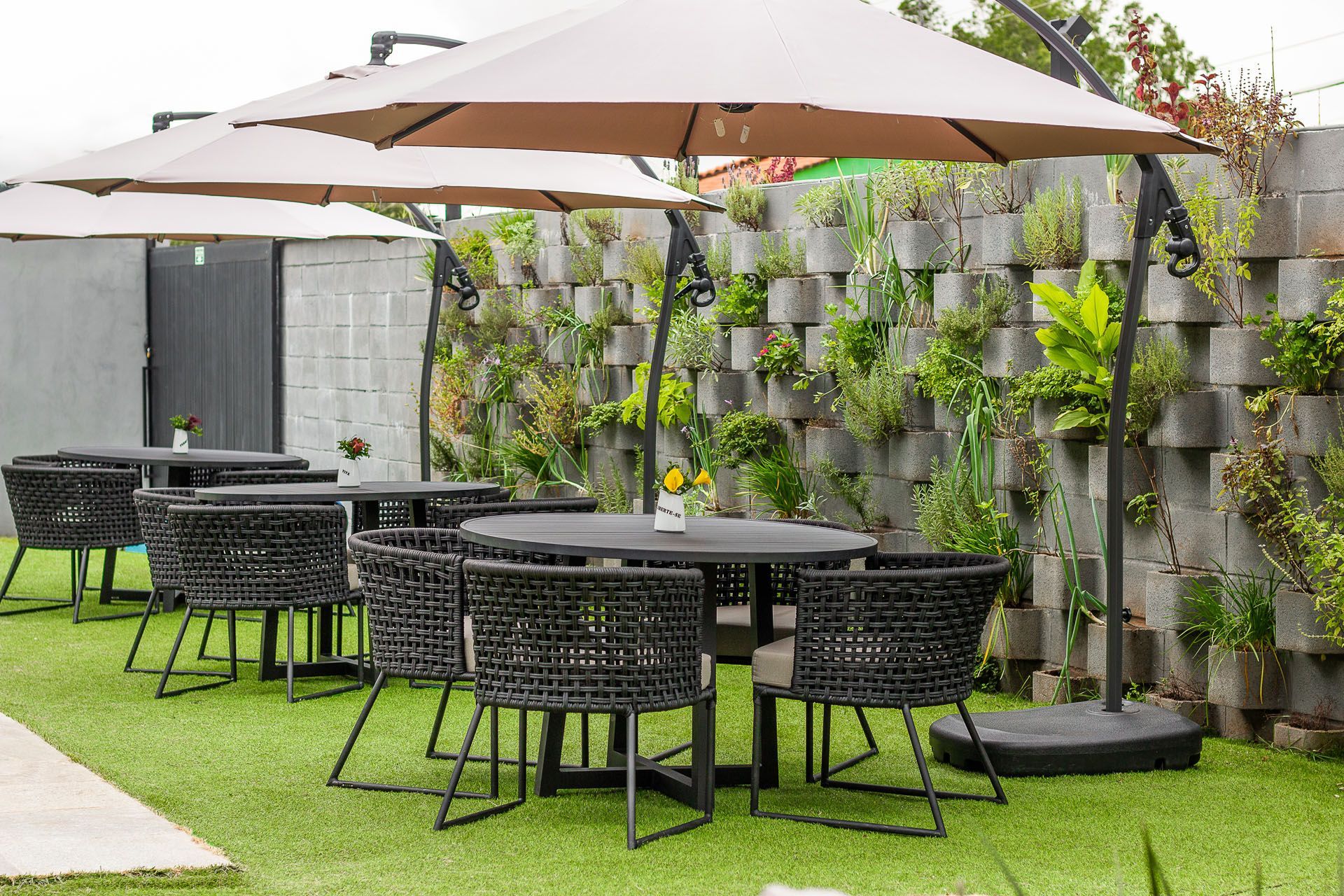 Área de refeições ao ar livre com mesas, cadeiras e guarda-sóis, junto a uma parede com plantas.