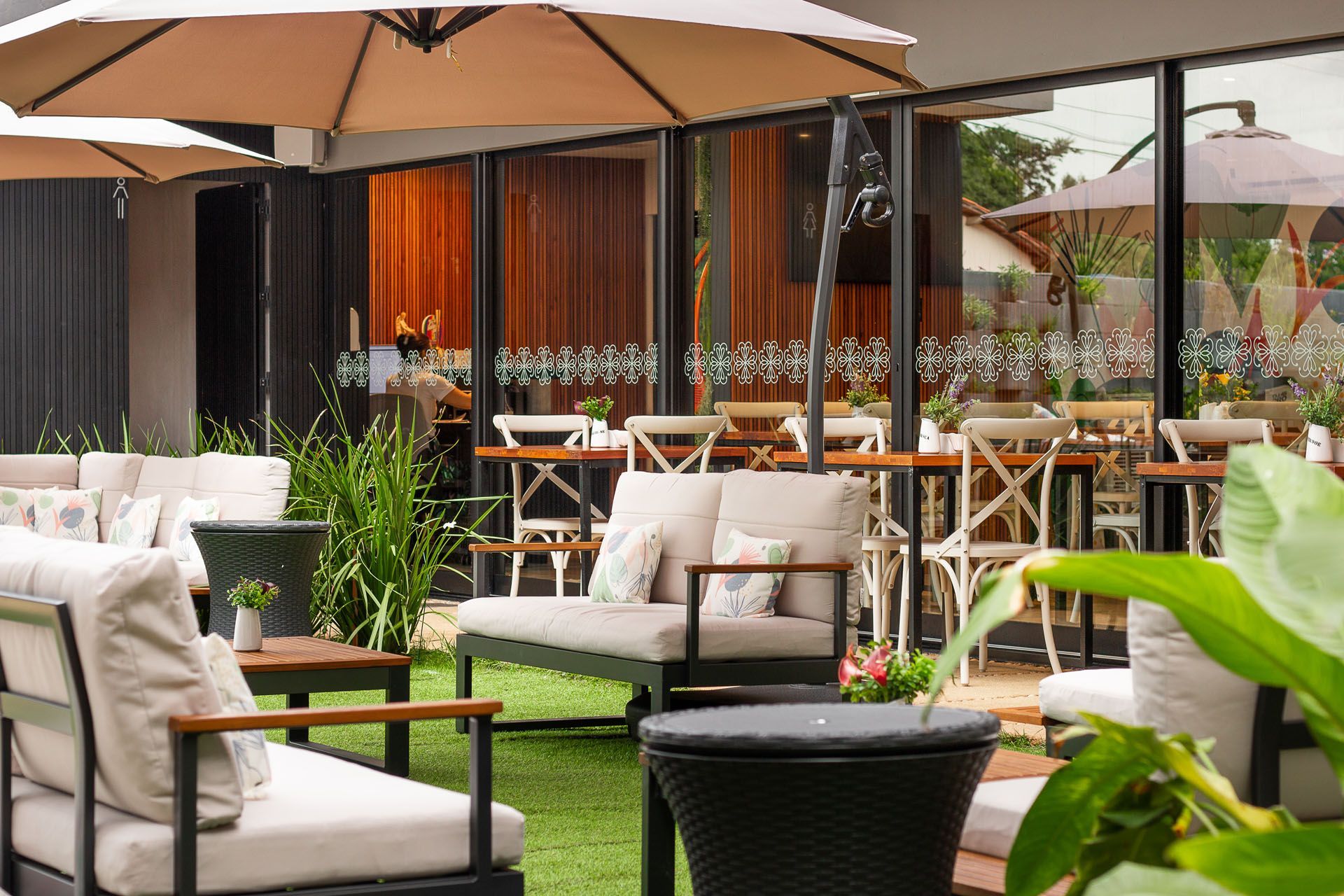 Pátio externo do café com mesas sob guarda-sóis e ao lado de janelas de vidro.