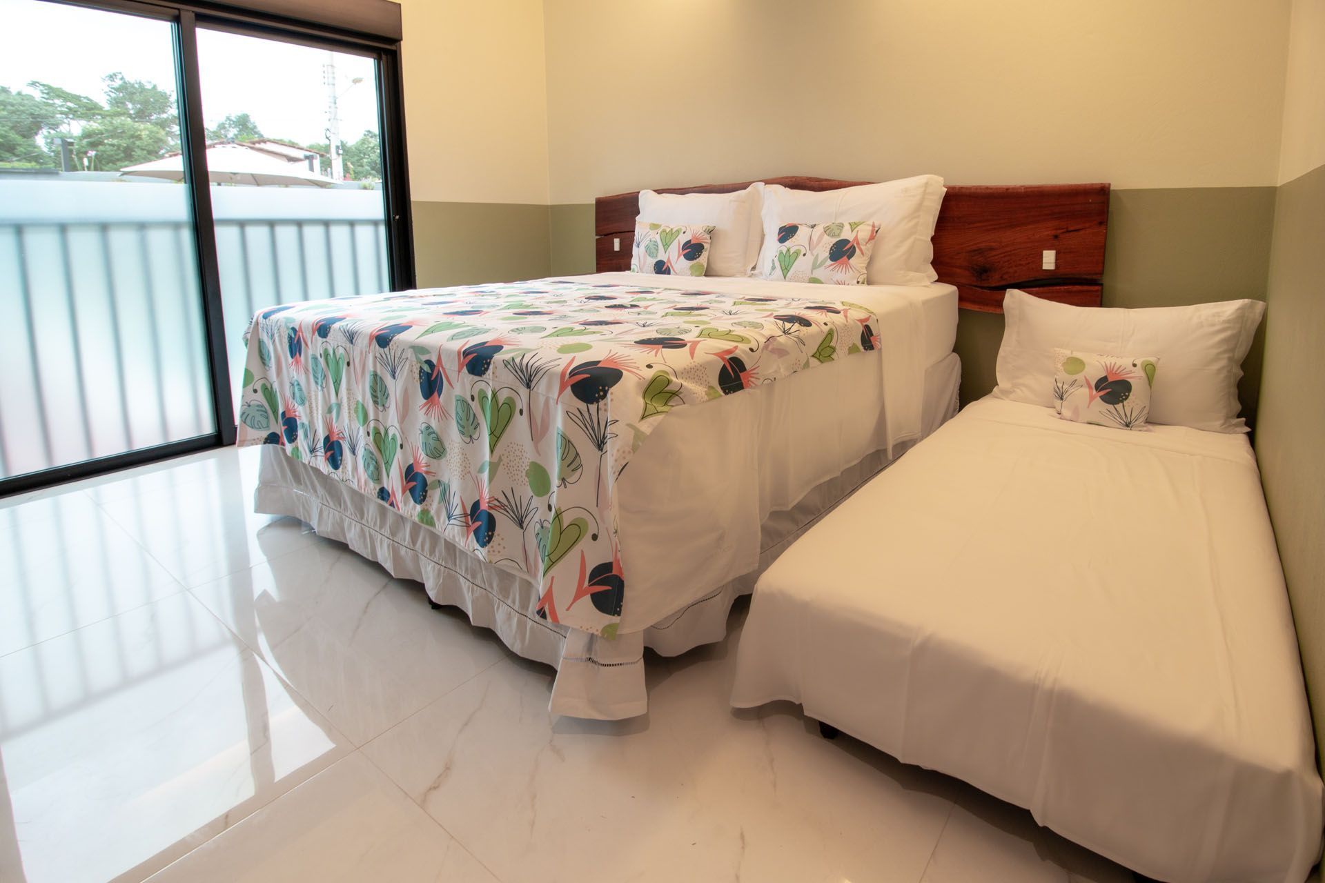 Quarto com uma cama de casal, uma cama de solteiro, roupa de cama branca e uma porta de vidro deslizante para uma varanda.