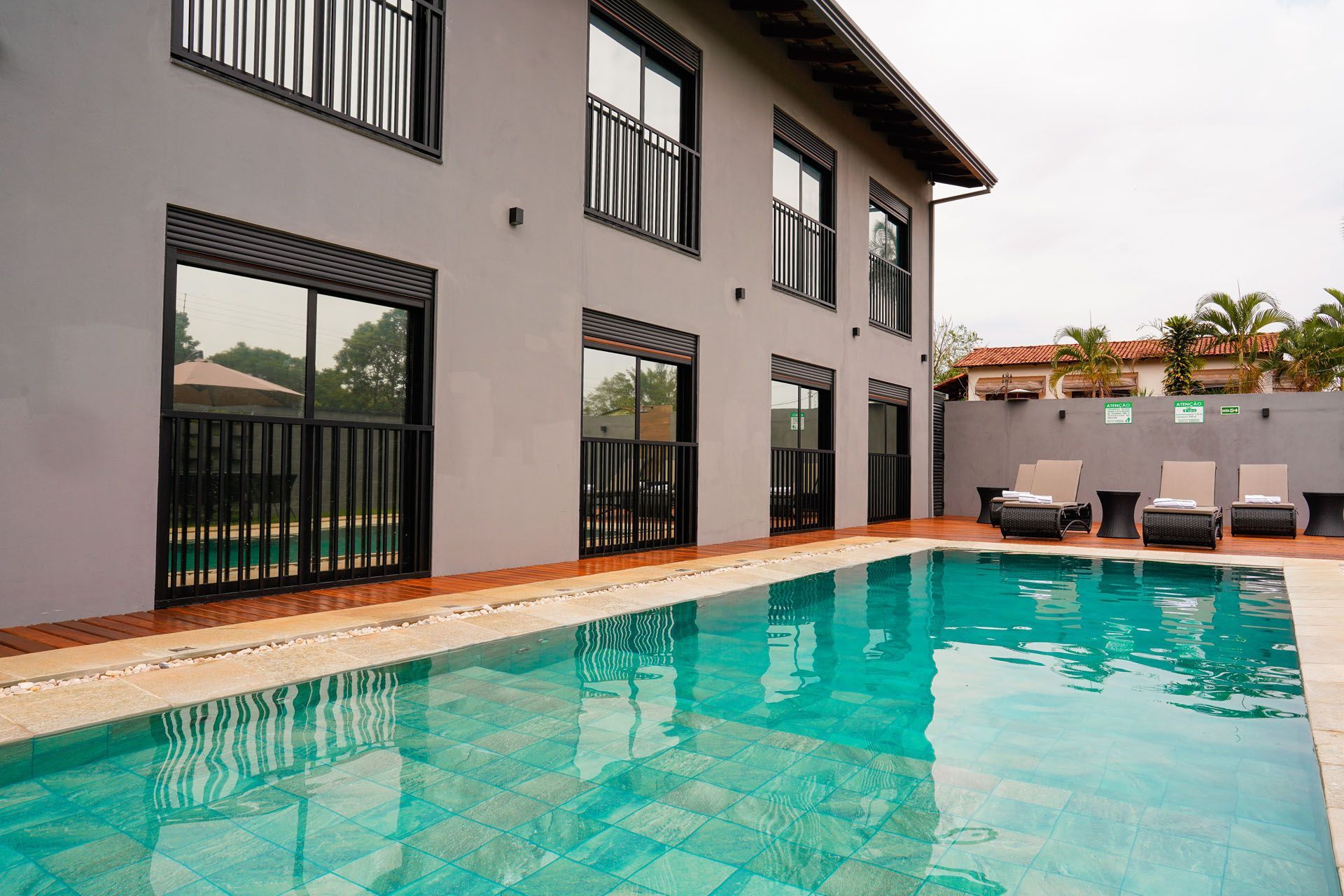 Prédio cinza de dois andares com janelas de moldura preta, com vista para uma piscina com espreguiçadeiras.